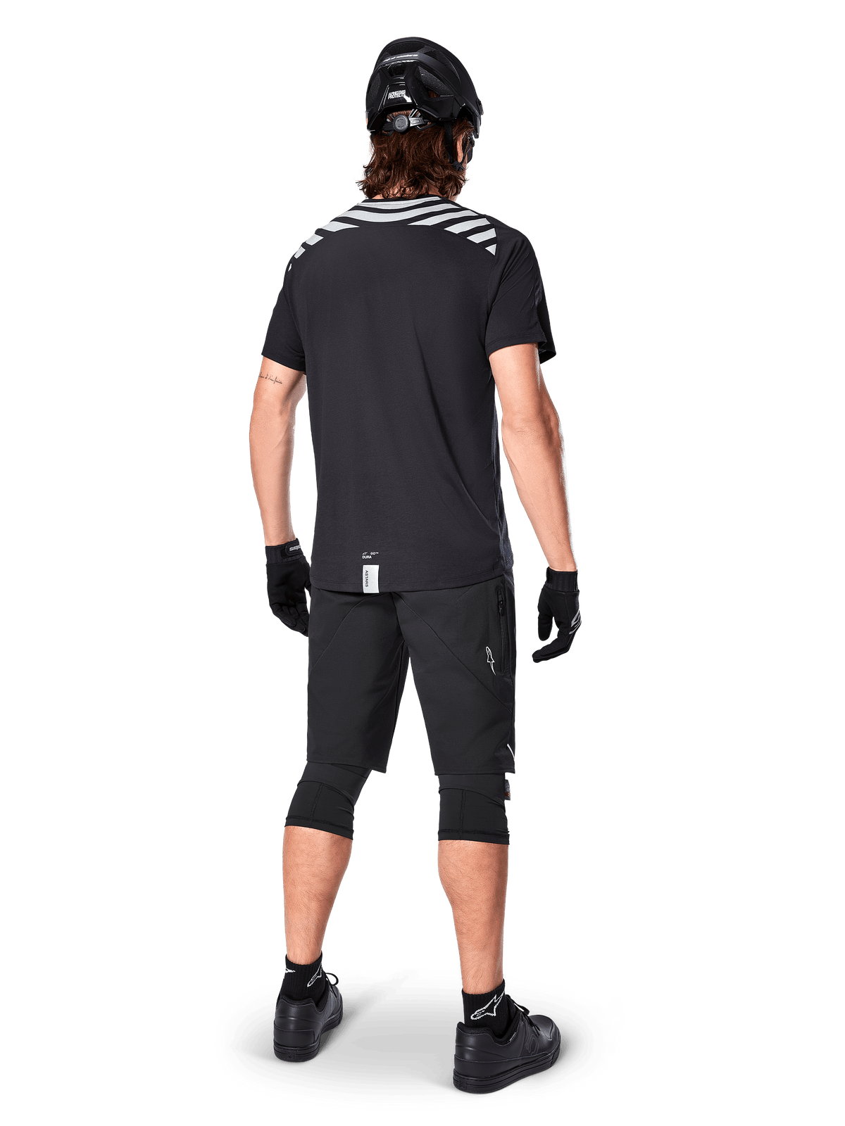 Maglia Alpinestars A-Dura Dri Oscar a maniche corte, nero riflettente, vista posteriore di un rider che indossa la maglia con pantaloncini da mountain bike neri, casco e ginocchiere, con grafiche riflettenti sulle spalle e design a coda lunga per MTB.