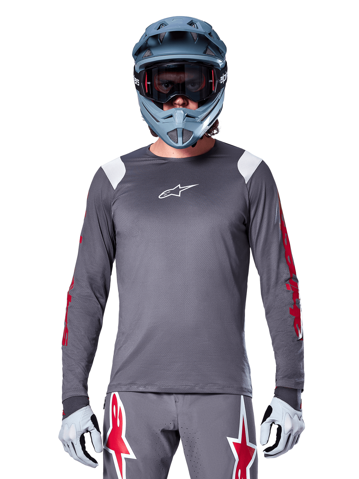 Ciclista MTB con maglia a maniche lunghe Alpinestars A-Supra Astar, grigio scuro con grafiche rosse sulle maniche e dettagli bianchi sulle spalle, pantaloni grigi coordinati con loghi a stella rossi, casco integrale blu e maschera, equipaggiamento da ciclismo fuoristrada coordinato