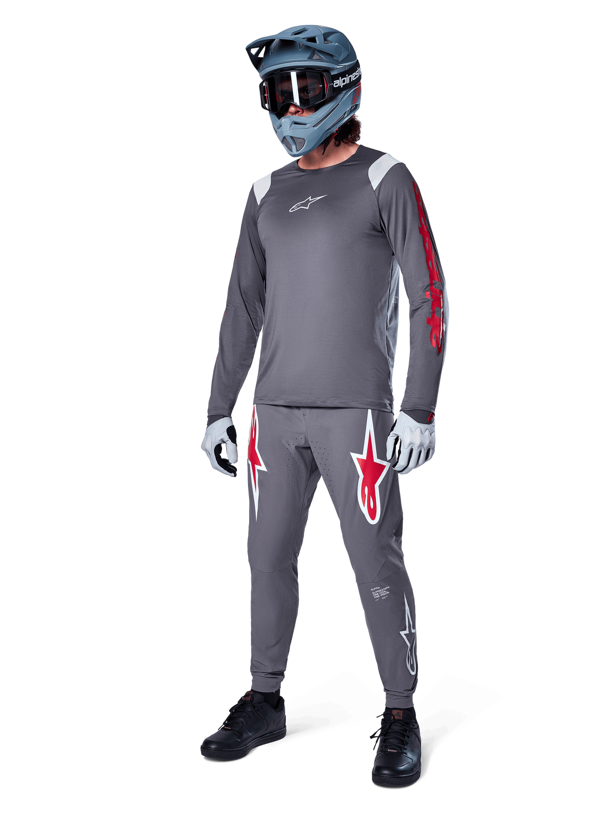 Maglia a maniche lunghe Alpinestars A-Supra Astar in grigio scuro, con branding rosso sulle maniche e logo bianco sul petto, indossata da un rider con pantaloni grigi coordinati, casco integrale grigio-blu e guanti tecnici, attrezzatura coordinata da mountain bike per il gravity.