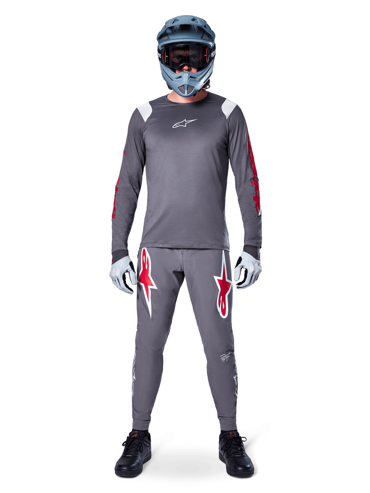 Ciclista MTB con maglia a maniche lunghe Alpinestars A-Supra Astar, grigio scuro con accenti rossi e logo bianco sul petto, pantaloni grigi coordinati con grafiche a stella rosse, casco integrale blu con maschera, guanti grigio chiaro e scarpe nere, completo tecnico professionale da mountain bike