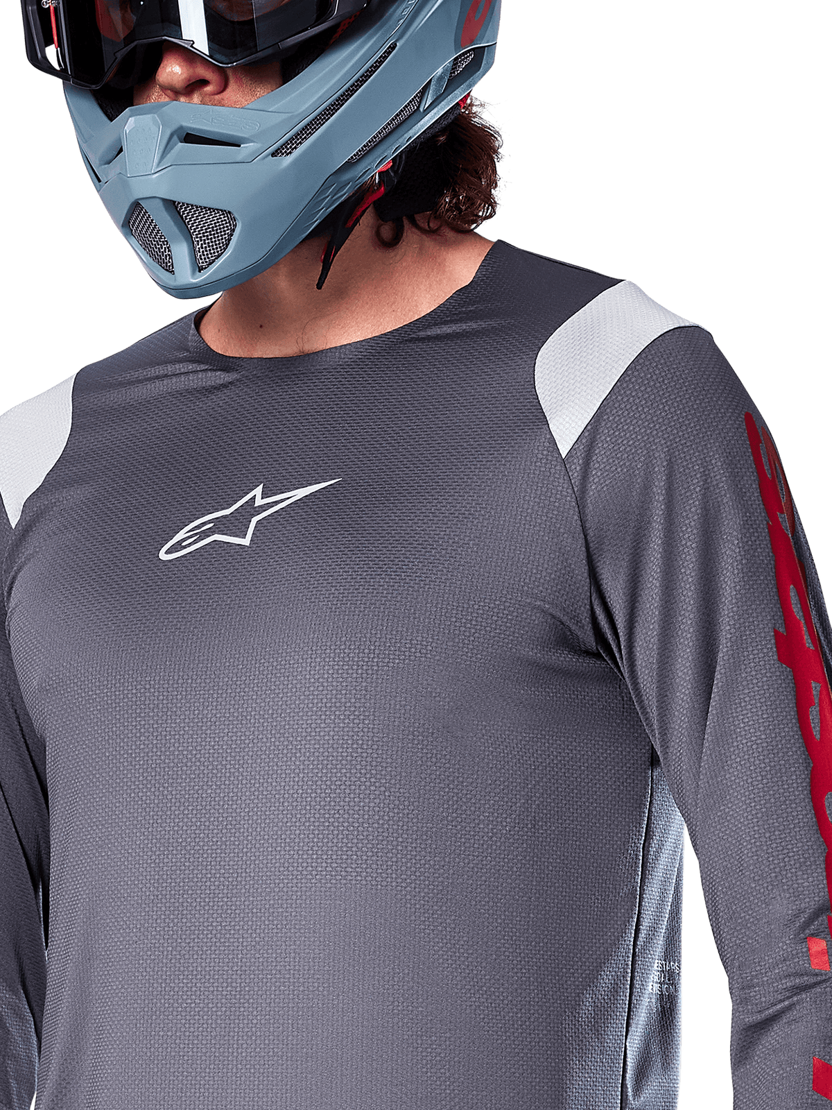 Alpinestars A-Supra Astar Jersey a maniche lunghe, grigio scuro con inserti bianchi sulle spalle e grafiche rosse sulle maniche, indossata con un casco integrale grigio-azzurro chiaro, abbigliamento tecnico per mountain bike