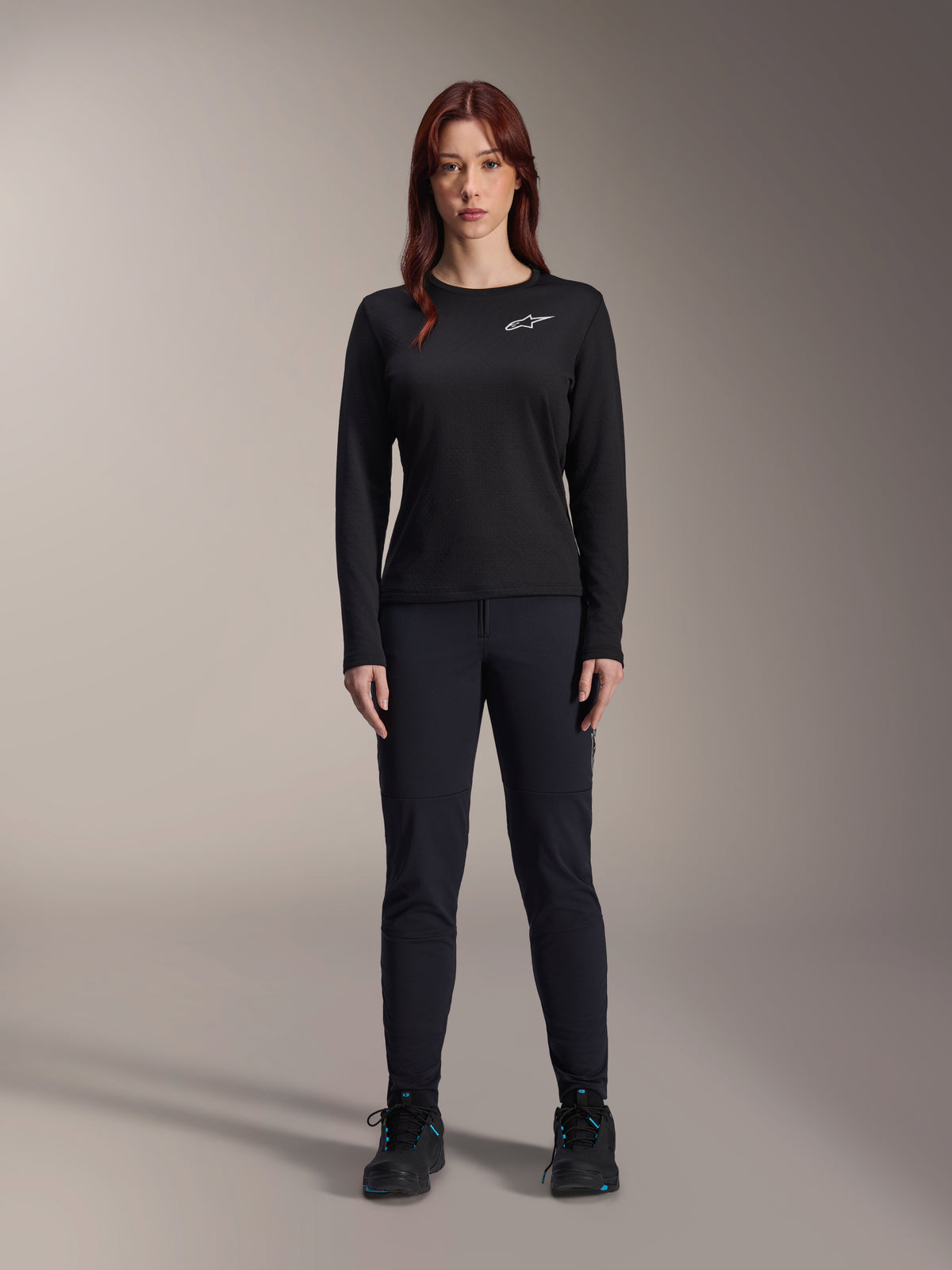 Alpinestars Stella A-Dura Thermal Jersey, maglia da mountain bike a maniche lunghe da donna in nero, con logo bianco sul petto, indossata con abbigliamento nero coordinato e scarpe da ciclismo, ritratto in studio.