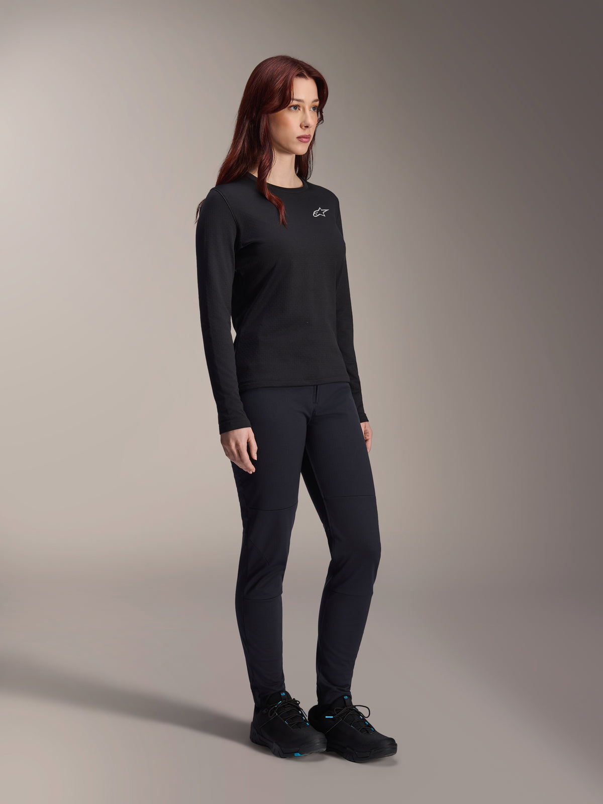 Donna che indossa la maglia Alpinestars Stella A-Dura Thermal Jersey, maglia MTB nera a maniche lunghe con logo bianco sul petto, abbinata a pantaloni neri coordinati e scarpe sportive, ritratto in studio per abbigliamento tecnico da ciclismo