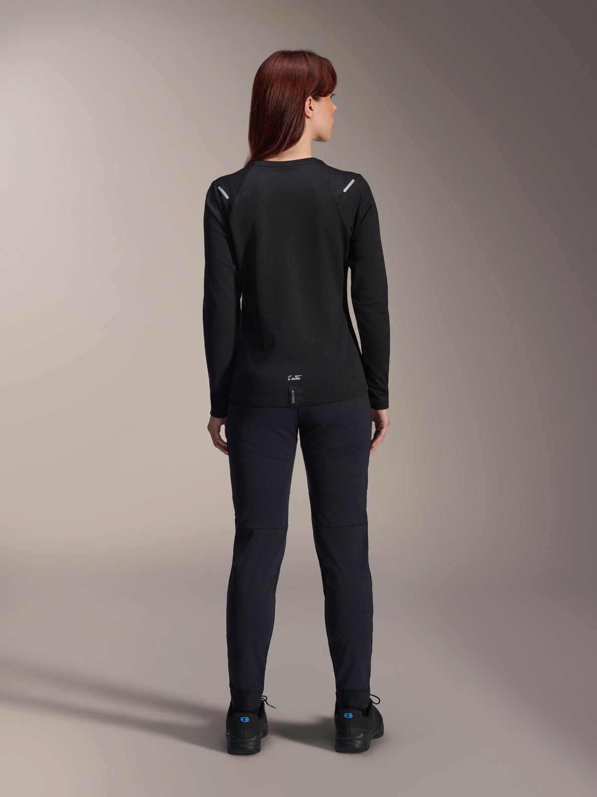 Alpinestars Stella A-Dura Thermal Jersey da donna, vista posteriore, nero, maglia da ciclismo a maniche lunghe con pile isolante, indossata con pantaloni e scarpe neri coordinati, caratterizzata dal piccolo logo A-Dura sulla parte bassa della schiena e dettagli riflettenti sulle spalle, specifica per MTB.