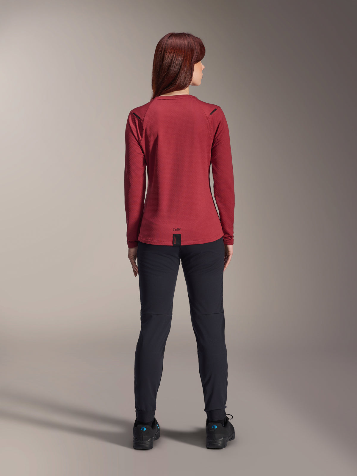 Donna che indossa la maglia termica Alpinestars Stella A-Dura, bordeaux, vista posteriore, maglia da ciclismo isolante a maniche lunghe con coda posteriore allungata, abbinata a pantaloni neri e scarpe da ciclismo nere, abbigliamento da mountain bike.