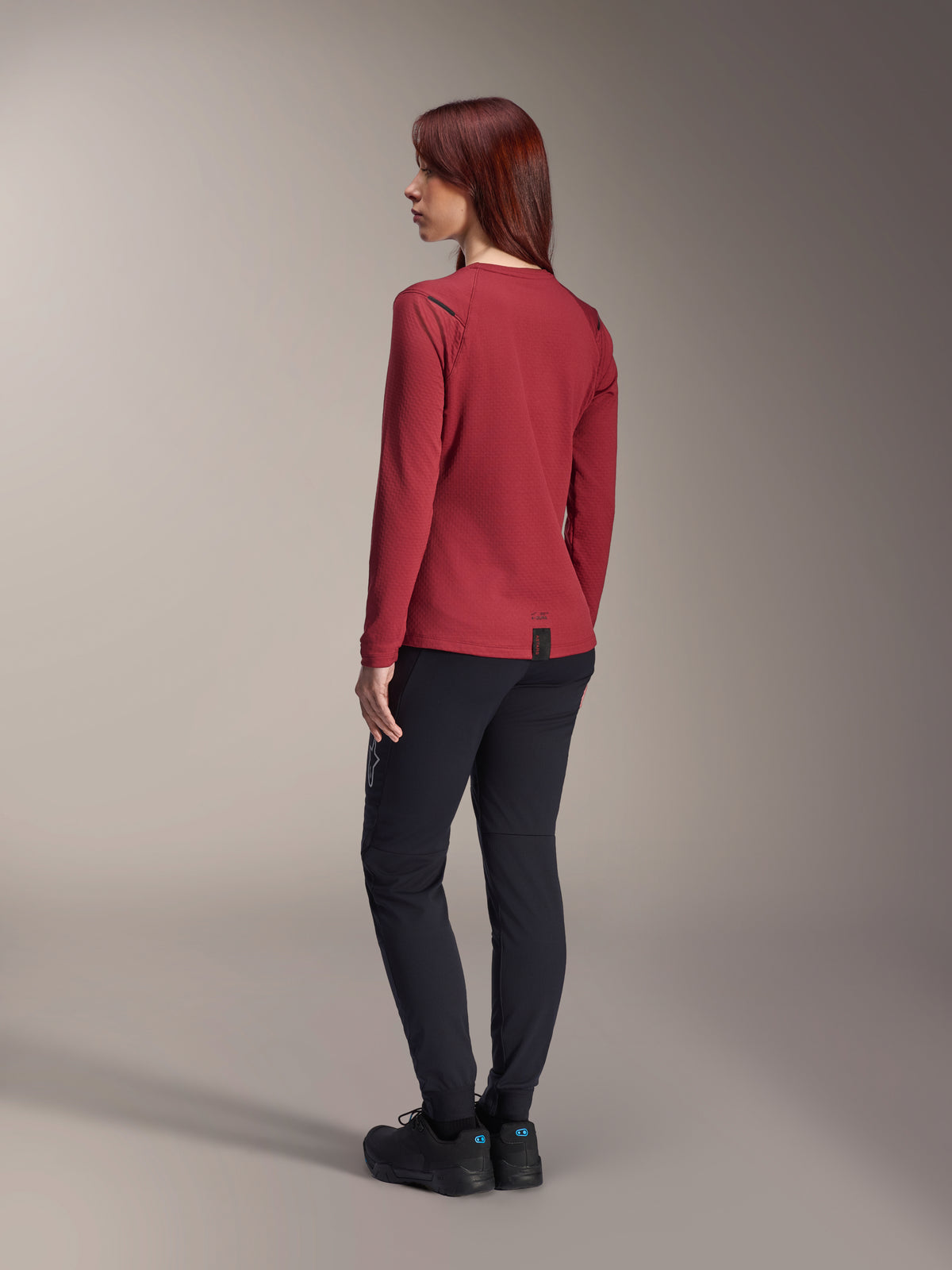 Alpinestars Stella A-Dura Thermal Jersey, maglia da mountain bike a maniche lunghe da donna, bordeaux, vista posteriore che evidenzia la coda allungata e la trama del tessuto termico, indossata con pantaloni da MTB neri e calzature nere