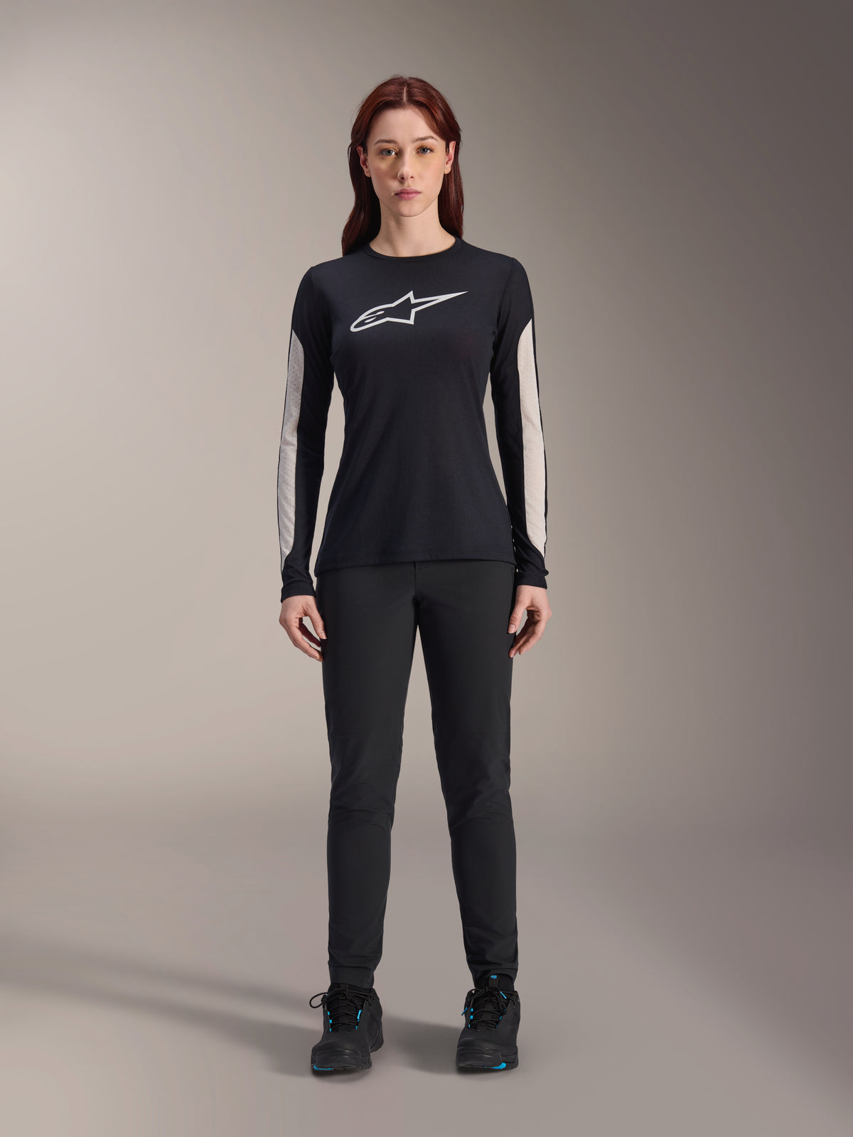 Donna che indossa la maglia a maniche lunghe Alpinestars Stella A-Dura Dri Astar, nera con logo bianco sul petto e pannelli delle maniche in mesh chiaro, abbinata a pantaloni neri e scarpe nere, abbigliamento tecnico da mountain bike