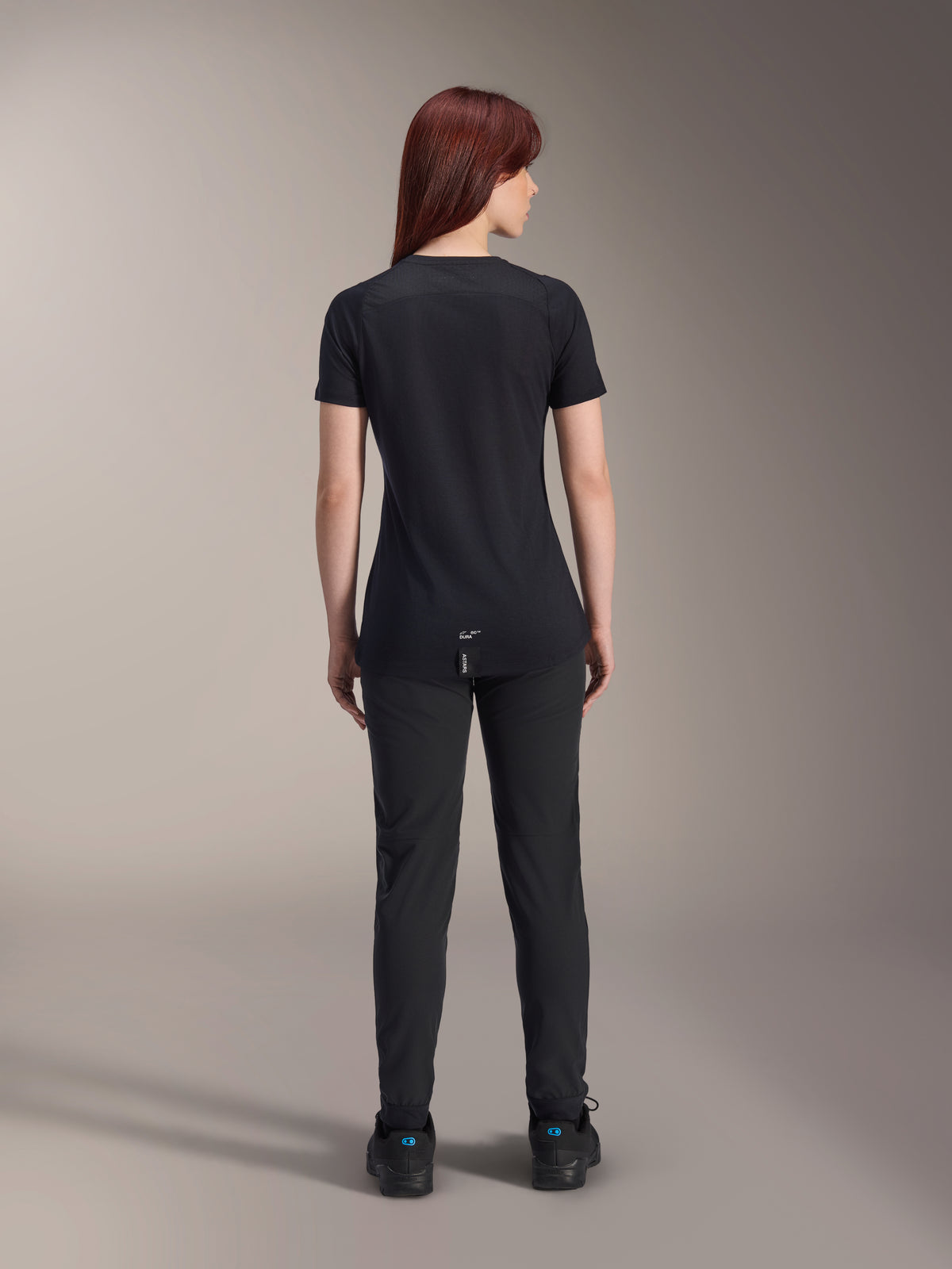 Alpinestars Stella A-Dura Dri Switch Short Sleeve Jersey da donna, nero, vista posteriore su una modella, con coda posteriore più lunga e pannelli in mesh tecnico, abbinata a pantaloni neri, abbigliamento tecnico da ciclismo MTB