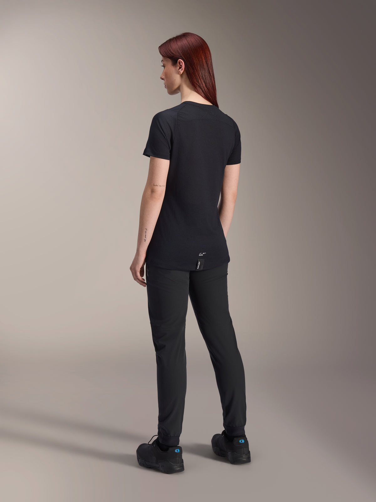 Modella con i capelli rossi che indossa la maglia Alpinestars Stella A-Dura Dri Switch a maniche corte in nero, vista posteriore, abbinata a pantaloni e scarpe da mountain bike neri, che mostra la vestibilità tecnica e la coda più lunga per la copertura durante la pedalata.