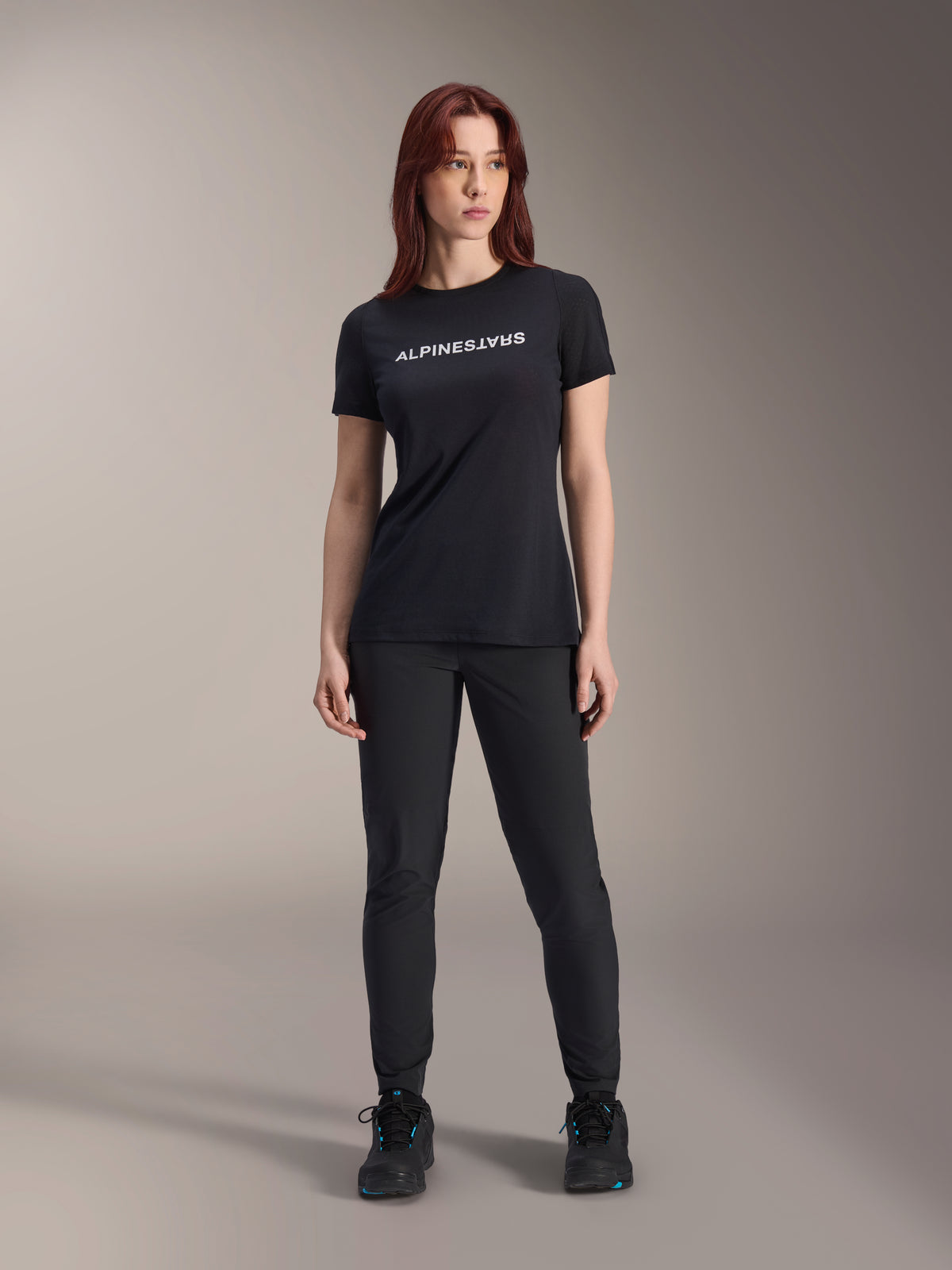 Modella con maglia a maniche corte Alpinestars Stella A-Dura Dri Switch da donna in nero, logo bianco sul petto, pantaloni tecnici neri e scarpe sportive nere, scatto in studio a figura intera
