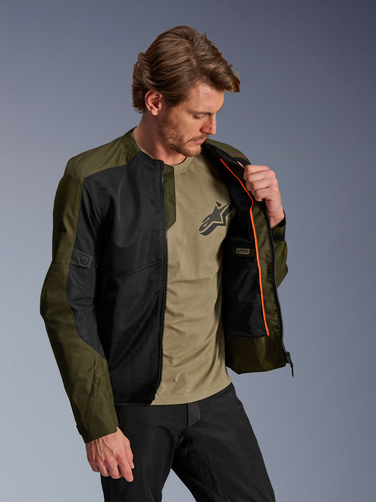 Giacca Alpinestars C-1 Air in Khaki e Nero, indossata da un modello per mostrare la fodera interna arancione e lo scomparto impermeabile, caratterizzata da ampi pannelli in rete tecnica per la ventilazione, stile urban contemporaneo abbinato a una t-shirt oliva e pantaloni neri