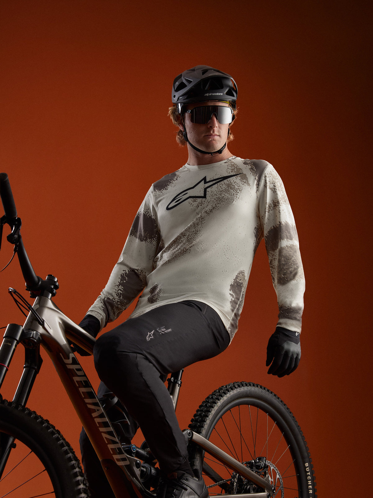 Maglia Alpinestars A-Dura Ride Sand Camo a maniche lunghe con logo sul petto, indossata da un rider su mountain bike.