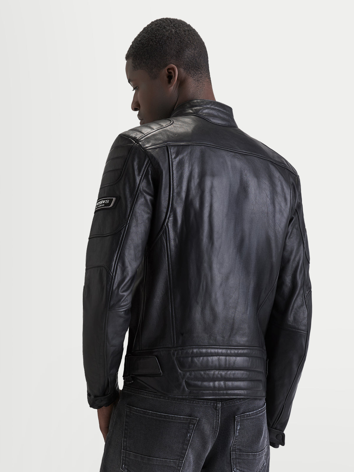 Alpinestars Revern Giacca di pelle, giacca da moto in pelle da uomo, colore nero, vista posteriore su un modello che mostra la struttura in pelle bovina pieno fiore, collo da motociclista classico e dettagli con marchio in stile heritage