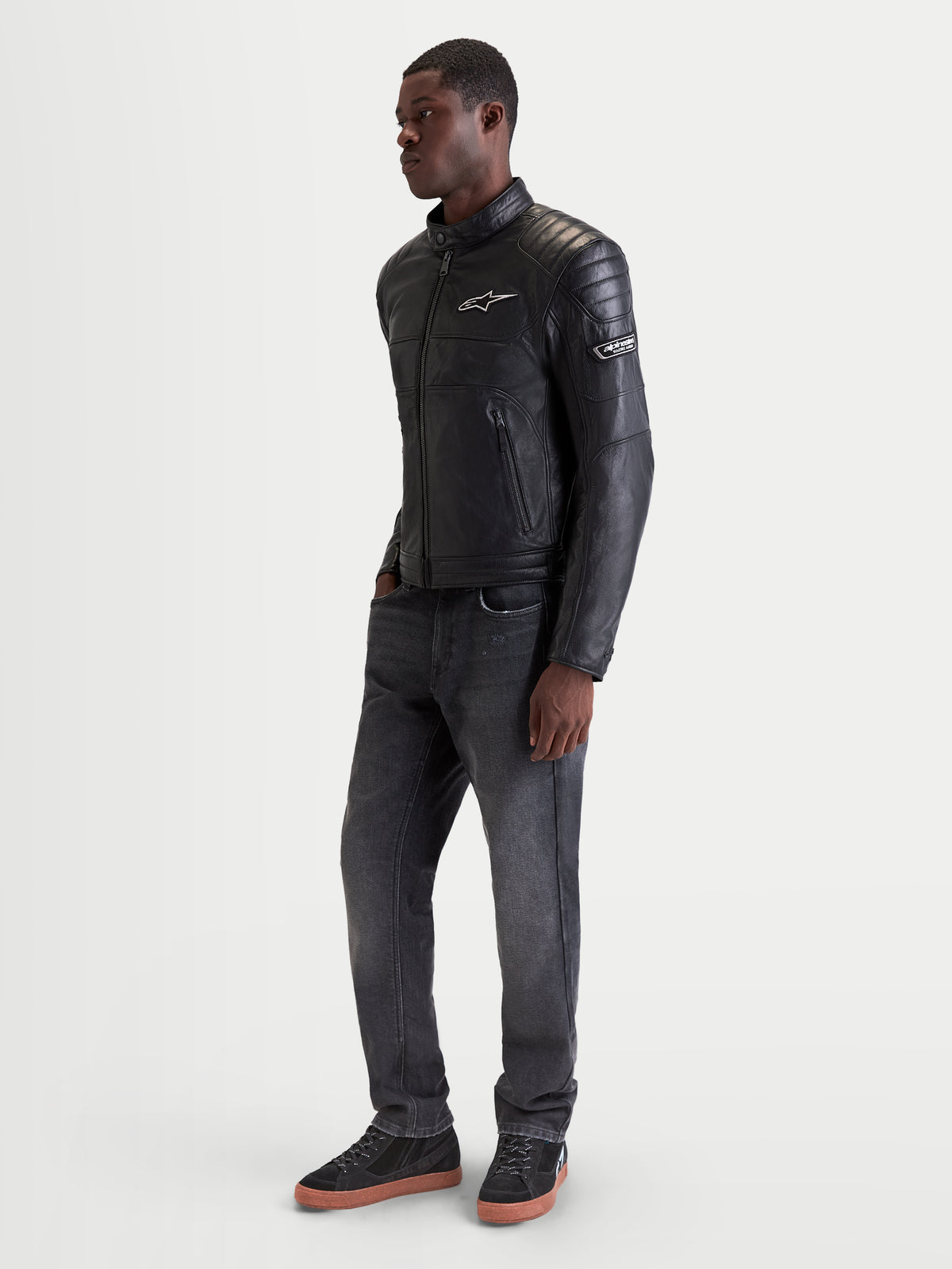 Alpinestars Revern Giacca in pelle, giacca da moto urbana da uomo in colore nero, vista laterale indossata da un modello che mostra lo stile heritage e la struttura in pelle bovina pieno fiore