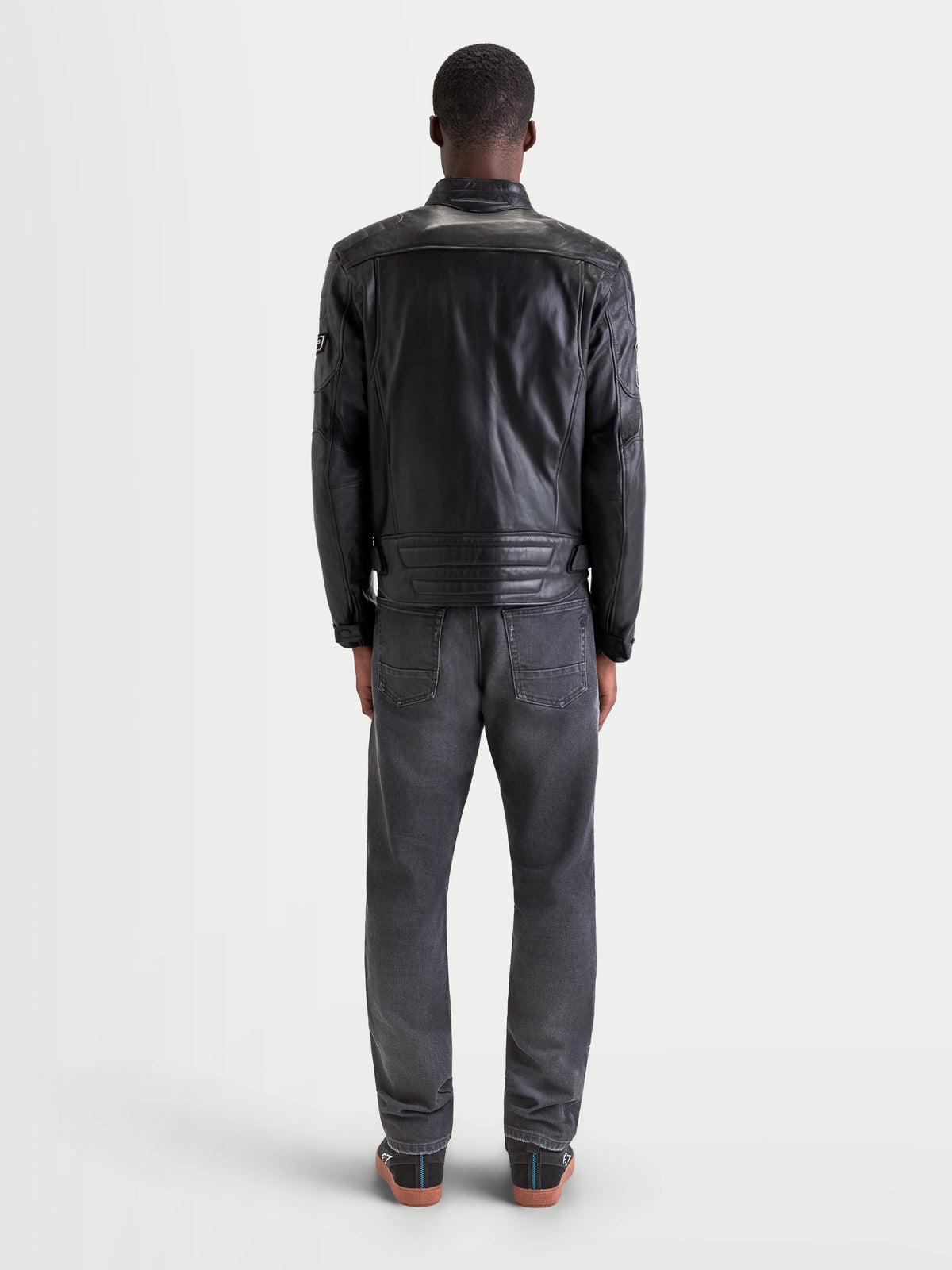 Alpinestars Revern Giacca di pelle, giacca da moto urbana contemporanea da uomo in pelle nera, vista posteriore su un modello con jeans e scarpe da moto casual, che mette in risalto la struttura in pelle bovina pieno fiore e lo stile heritage da moto