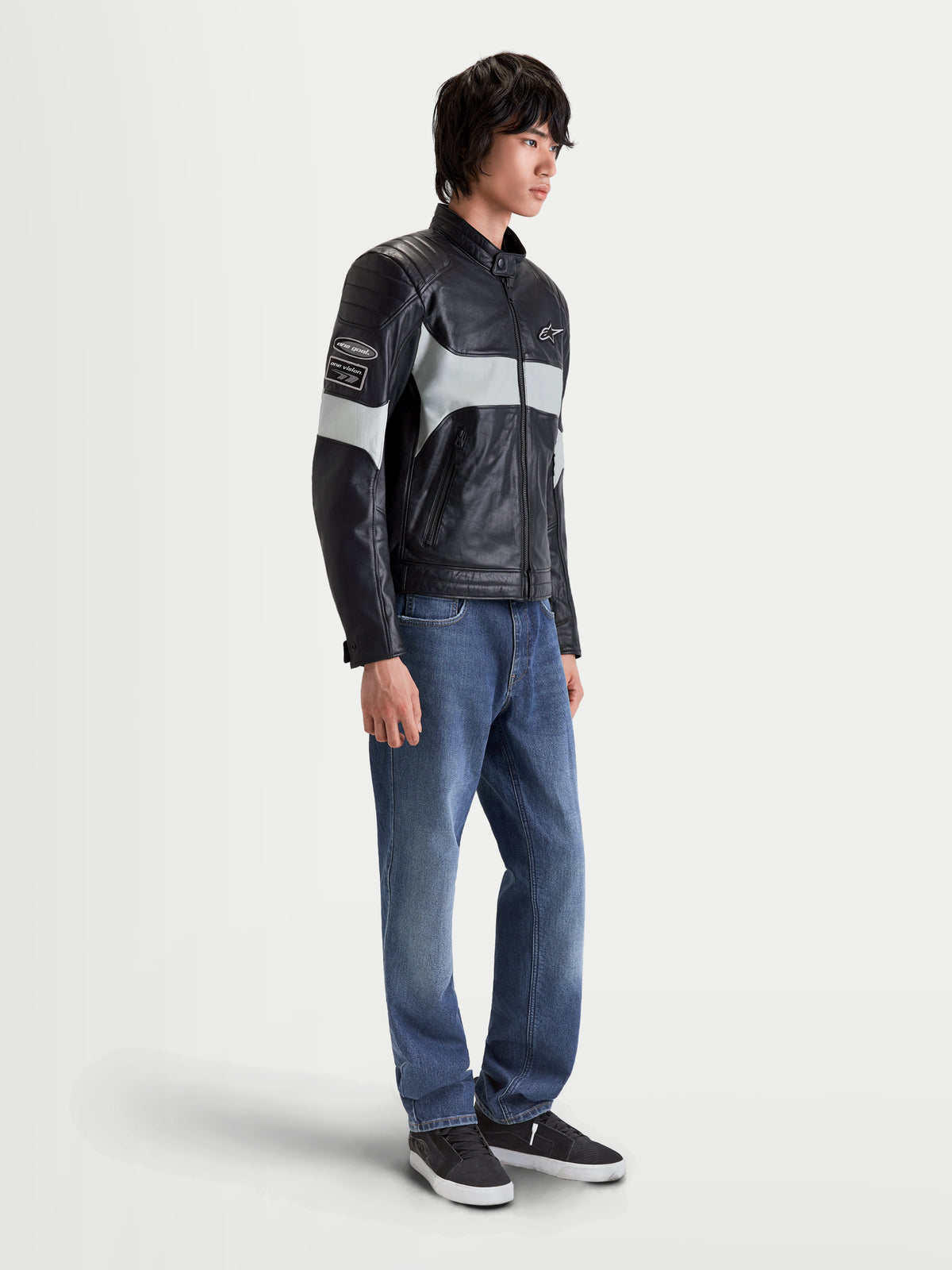 Alpinestars Revern Giacca di pelle, giacca da moto urbana in pelle da uomo nera e grigio metallizzato, vista laterale indossata da un modello con jeans, caratterizzata da struttura in pelle pieno fiore in stile heritage