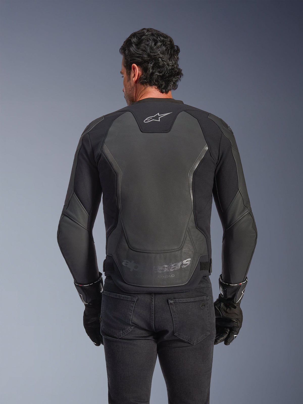 Giacca in pelle Alpinestars GP Force V2, nera, vista posteriore di un uomo che indossa la giacca con guanti neri e jeans, con ampi pannelli protettivi in pelle sulla schiena, logo Alpinestars sulla parte superiore e marchio in rilievo sulla parte bassa della vita, abbigliamento da moto sportivo