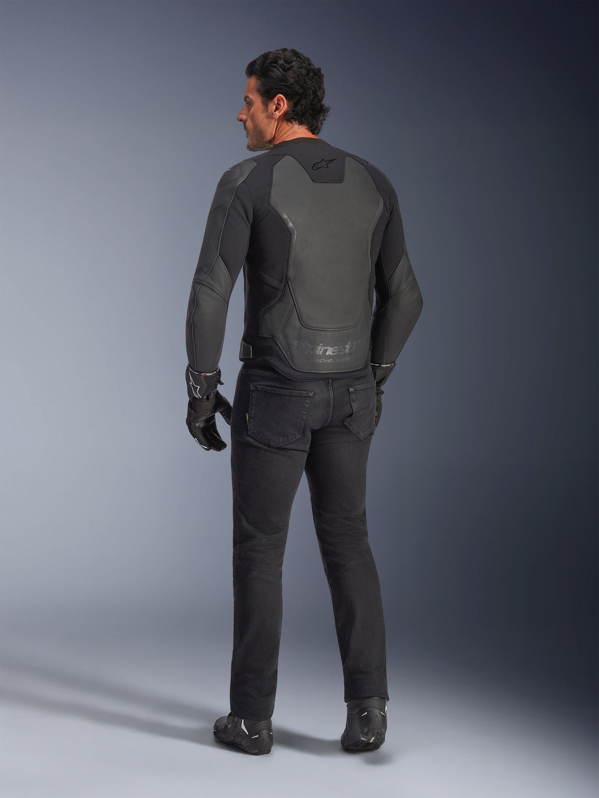 Vista posteriore di un modello che indossa la giacca in pelle Alpinestars GP Force V2 in Black Black, caratterizzata da una costruzione in pelle multi-pannello e vestibilità sportiva, abbinata a pantaloni, guanti e stivali da moto neri per un look completo ispirato al racing.