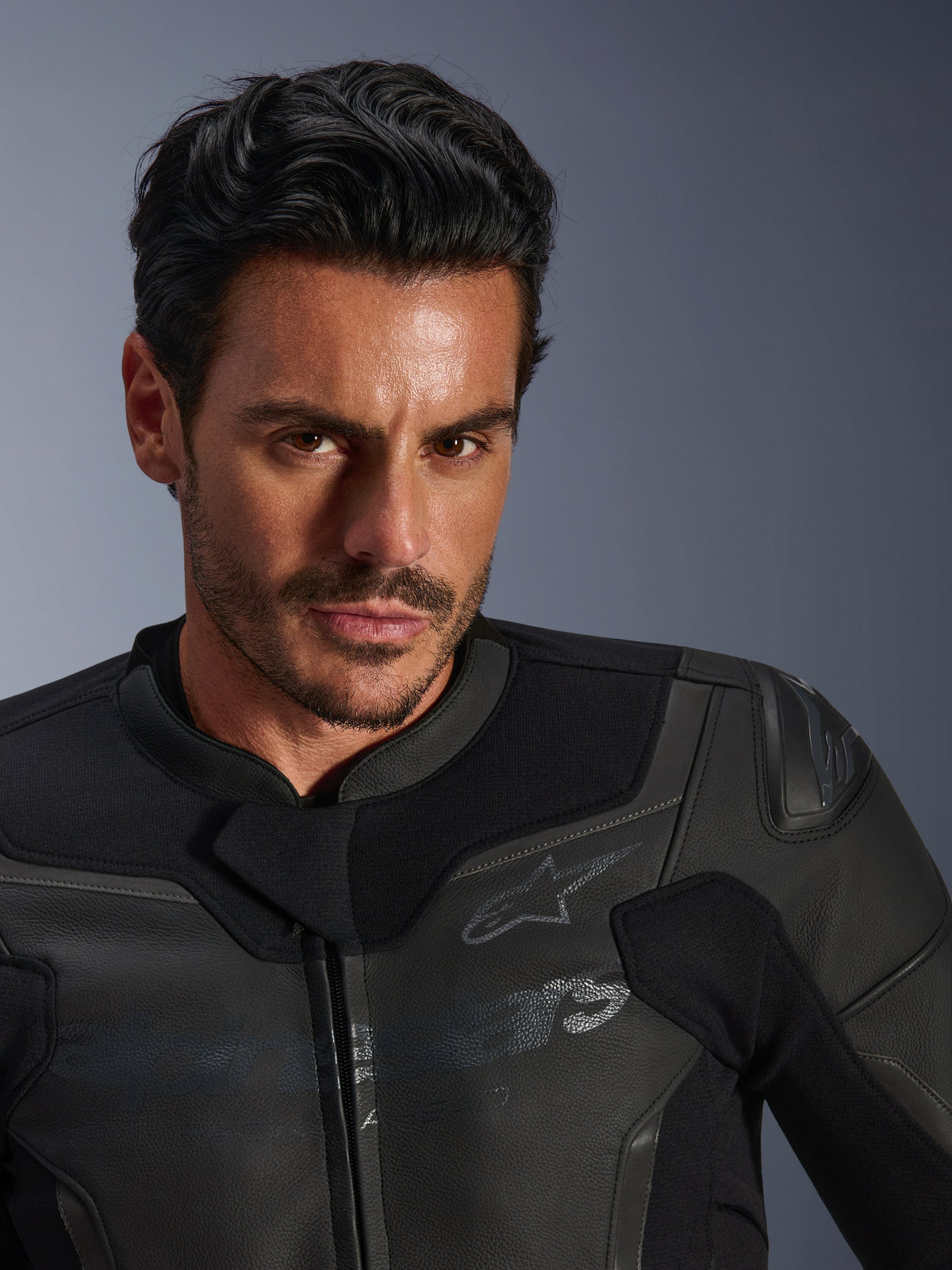 Ritratto in primo piano di un modello maschile che indossa la giacca in pelle Alpinestars GP Force V2, nera, caratterizzata da una costruzione in pregiata pelle bovina, pannelli elasticizzati in tessuto e un discreto logo a stella Alpinestars, design sportivo ed elegante per la guida su strada.