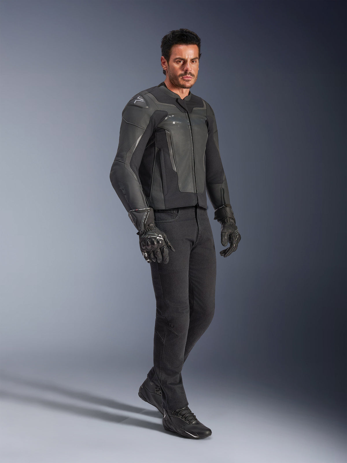 Uomo che indossa la giacca in pelle Alpinestars GP Force V2 in nero, abbinata a pantaloni neri e guanti da moto, scatto in studio a corpo intero, elegante costruzione in pelle bovina con protezioni integrate su spalle e gomiti, abbigliamento professionale da strada per moto sportiva