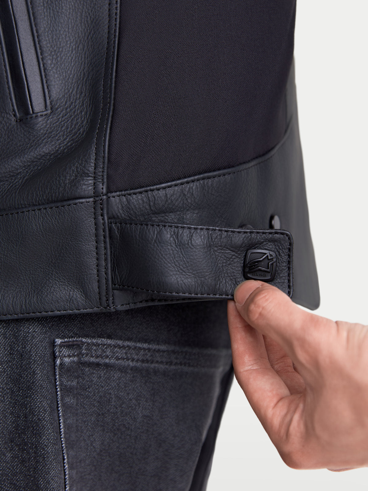 Alpinestars Maver X Giacca di pelle, giacca da moto urban da uomo, vista ravvicinata della linguetta di chiusura a scatto regolabile in vita in colore nero, pregiata struttura in pelle e inserti elasticizzati