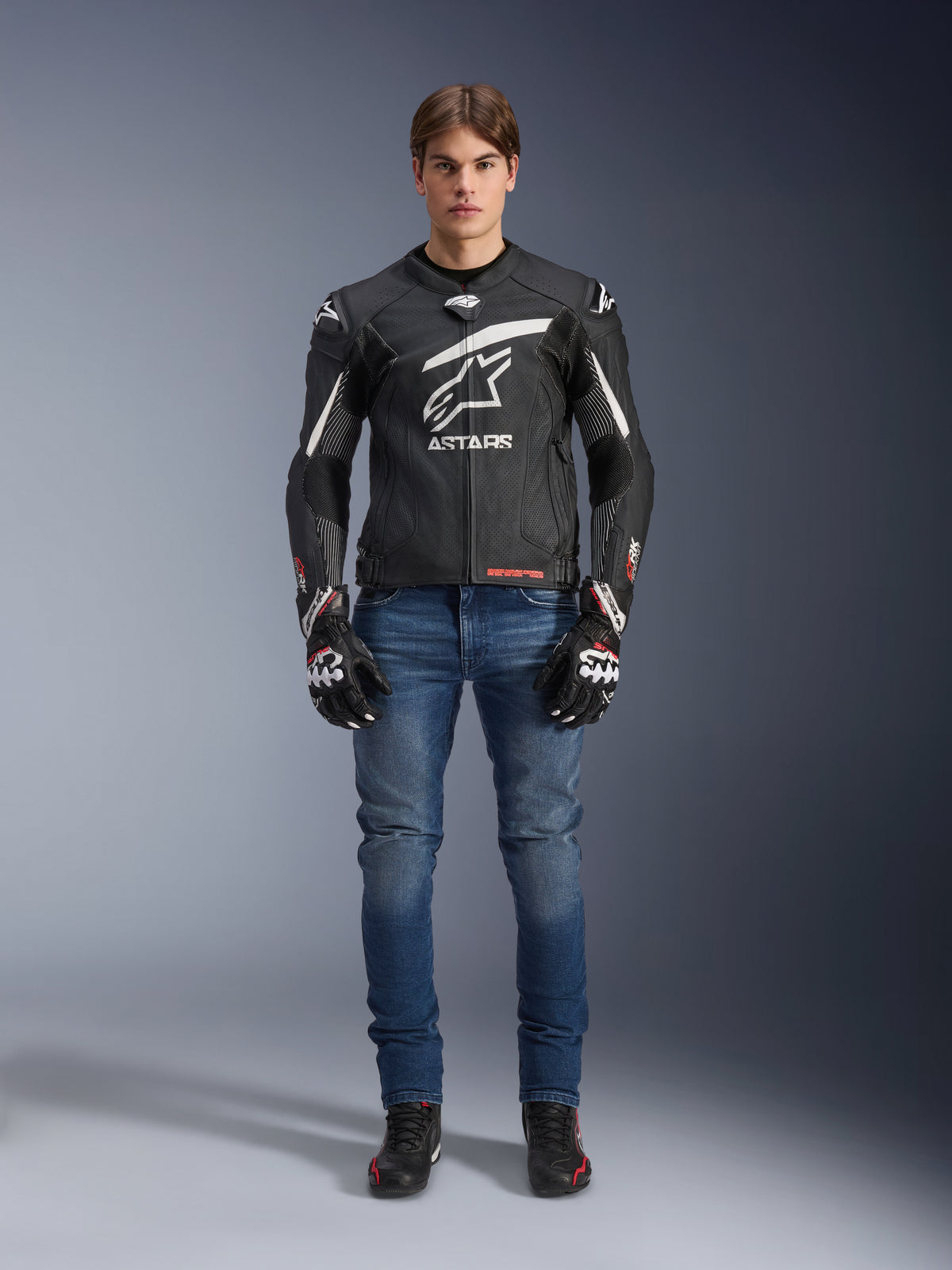 Modello che indossa la giacca in pelle Alpinestars GP Plus R V4 Rideknit in nero e bianco, abbinata a jeans da moto blu, guanti da moto neri e scarpe da guida nere, con un grande logo Astars bianco sul petto, pannelli in pelle traforata e strisce racing bianche sulle maniche, vista frontale a figura intera