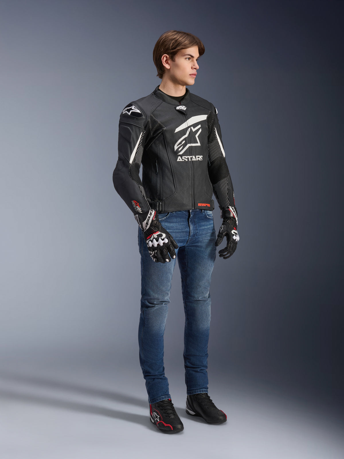 Giacca in pelle Alpinestars GP Plus R V4 Rideknit, nero e bianco, giacca da moto in pelle indossata da un modello con jeans blu e guanti neri, caratterizzata da un prominente logo Astars bianco sul petto, pannelli traforati e dettagli bianchi sulle spalle.