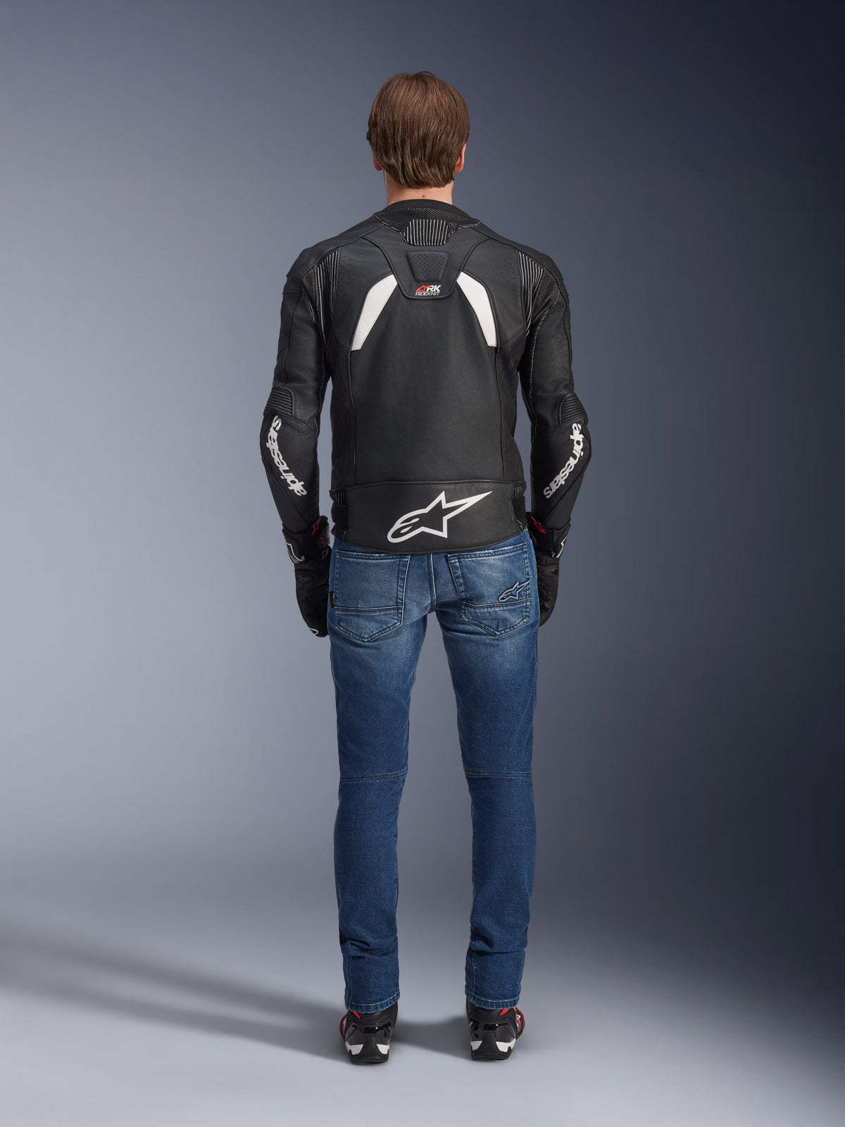 Vista posteriore della giacca in pelle Alpinestars GP Plus R V4 Rideknit, nero e bianco, con un grande logo bianco sulla parte bassa della schiena e branding sulle maniche, indossata con jeans da moto blu e stivali neri, evidenziando la costruzione tecnica e la vestibilità sportiva.