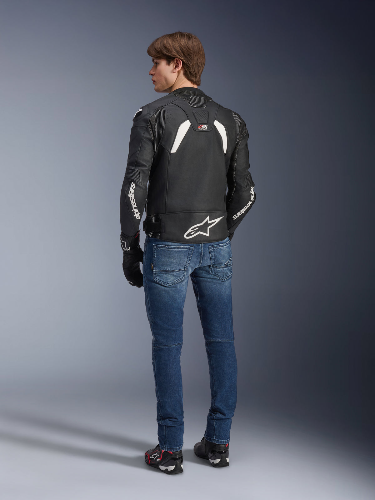 Giacca in pelle Alpinestars GP Plus R V4 Rideknit in nero e bianco, vista posteriore su un modello, con marchio Alpinestars bianco sulle maniche e un grande logo a stella sulla parte inferiore della schiena, indossata con jeans blu e scarpe da moto nere, abbigliamento tecnico da moto