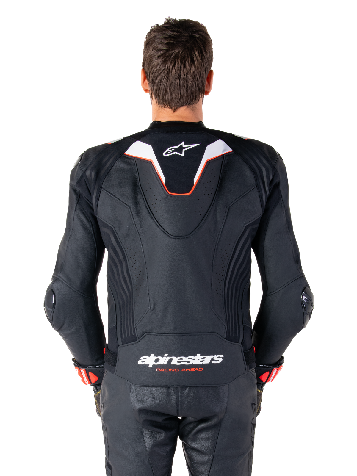 Vista posteriore di un pilota che indossa la giacca in pelle Alpinestars Missile V3 Ignition, in pelle nera con accenti bianchi e rossi, caratterizzata da una sottile gobba aerodinamica sportiva con logo bianco e marchio 'alpinestars RACING AHEAD' sulla parte bassa della schiena, abbinata a pantaloni da corsa e guanti in pelle nera coordinati.