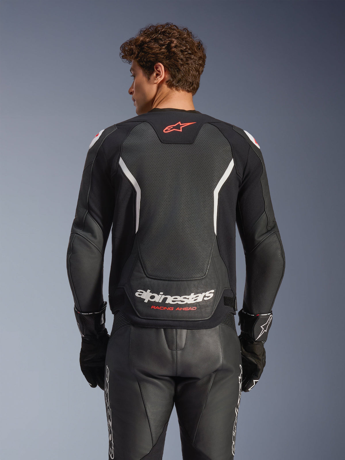 Giacca in pelle Alpinestars GP Force V2 Air, nero con accenti bianchi e rossi, vista posteriore con pannello posteriore in pelle traforata, branding Alpinestars bianco e dettagli del logo rossi, indossata con pantaloni racing e guanti in pelle nera coordinati.