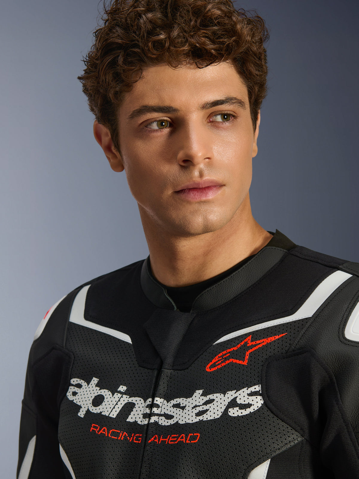 Ritratto di un modello maschile che indossa la giacca in pelle Alpinestars GP Force V2 Air in nero e bianco, caratterizzata da pelle traforata, logo bianco e scritta rossa 'RACING AHEAD' sul petto, abbigliamento sportivo da moto