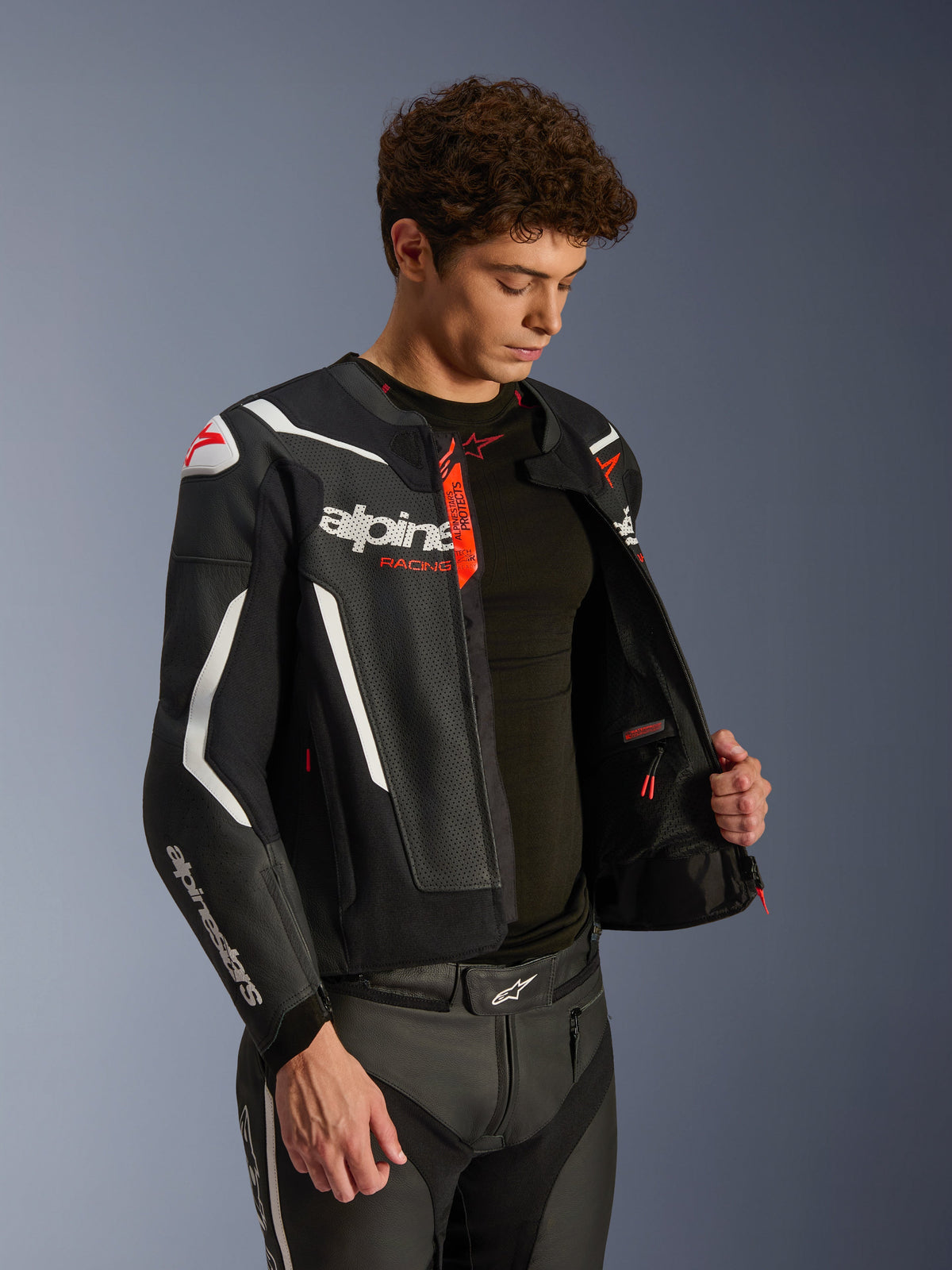 Giacca in pelle Alpinestars GP Force V2 Air, nera e bianca con accenti rossi, struttura in pelle traforata, aperta per mostrare l'interno e il base layer nero, abbinata a pantaloni in pelle coordinati, scatto professionale in studio per abbigliamento moto stradale