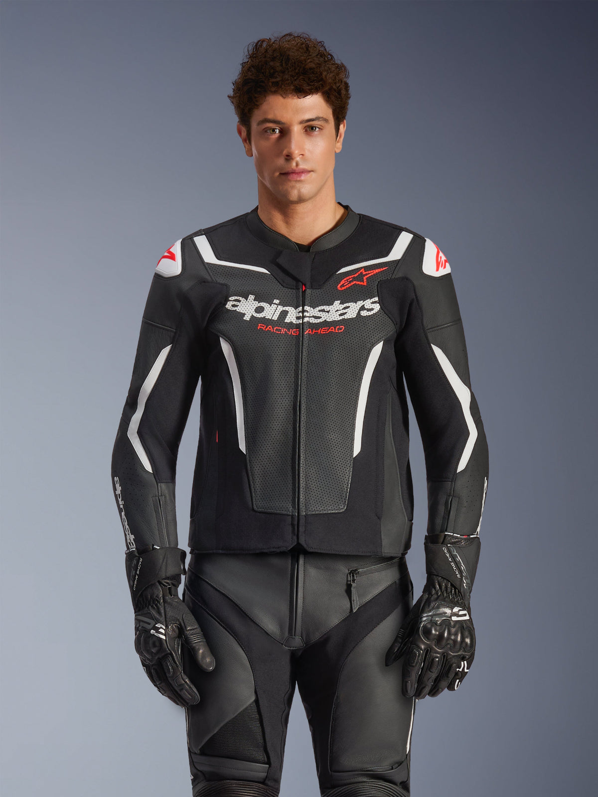 Giacca in pelle Alpinestars GP Force V2 Air, nero con dettagli bianchi e rossi, modello che indossa la giacca in pelle traforata con marchi 'alpinestars' e 'RACING AHEAD' sul petto, abbinata a pantaloni e guanti da corsa coordinati, abbigliamento sportivo premium da moto