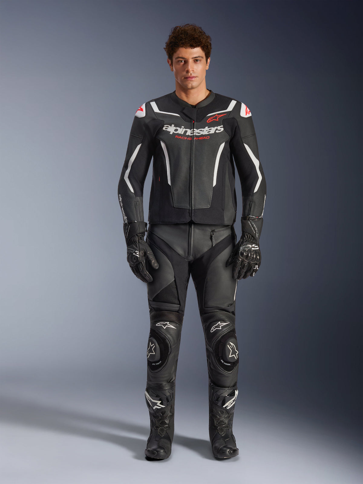 Modello maschile che indossa la giacca in pelle Alpinestars GP Force V2 Air, nera e bianca, abbinata a pantaloni in pelle, guanti e stivali coordinati, vista frontale sportiva, costruzione in pelle traforata con accenti bianchi e loghi Alpinestars