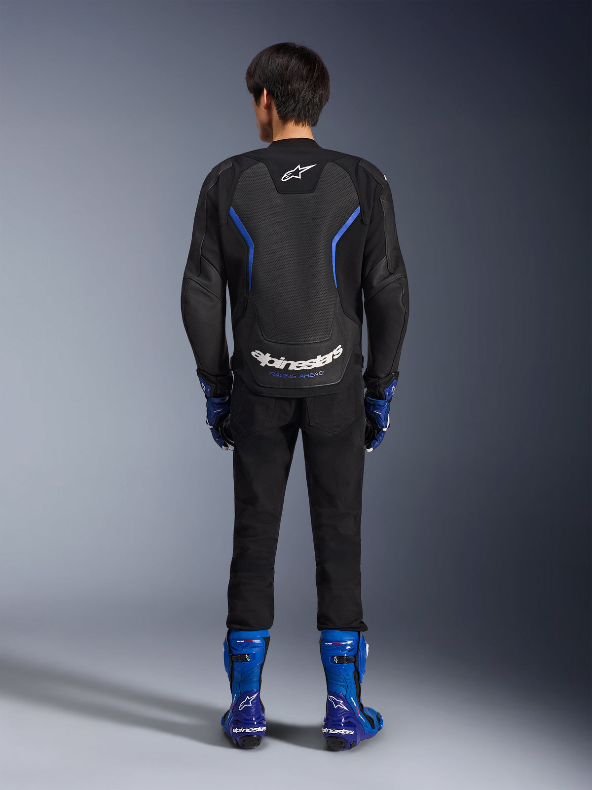 Giacca in pelle Alpinestars GP Force V2 Airflow, Black Metallic Blue, vista posteriore con pannelli in pelle traforata e logo bianco, indossata con stivali racing blu e pantaloni neri, abbigliamento sportivo da moto racing
