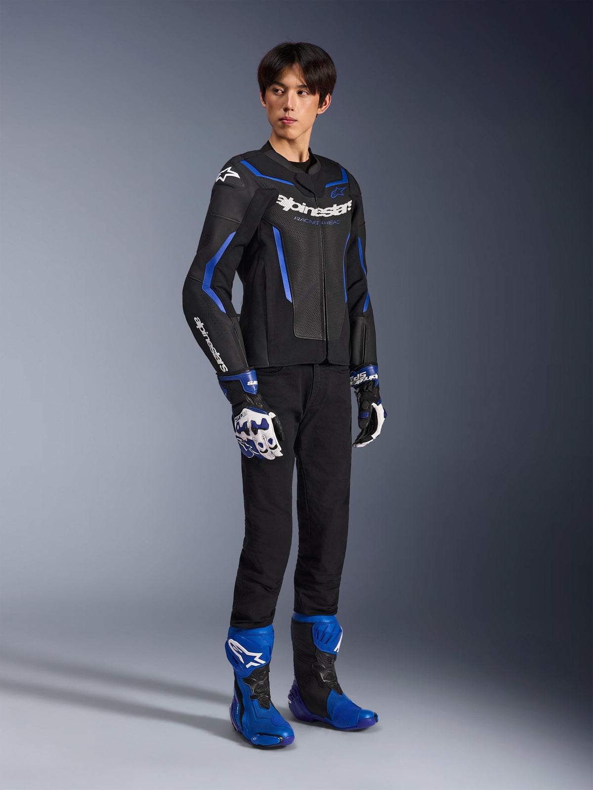 Pilota uomo che indossa la giacca in pelle GP Force V2 Airflow in Nero Blu Metallizzato, abbinata a guanti racing coordinati e stivali blu, con pelle traforata e loghi Alpinestars in evidenza, abbigliamento professionale da moto.