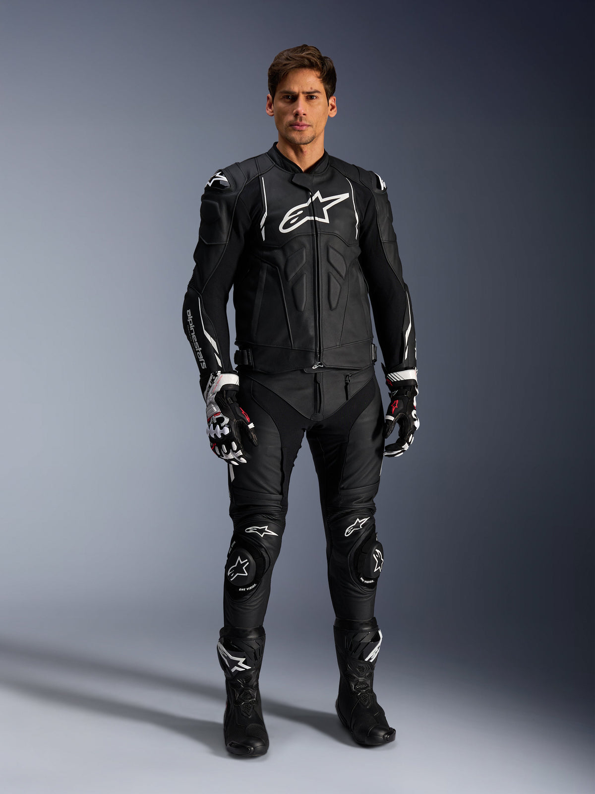 Alpinestars Missile V3 Leather Pants, neri, indossati da un pilota uomo come parte di un completo da corsa in pelle, con loghi Alpinestars bianchi, saponette protettive sulle ginocchia, stivali e guanti da gara.