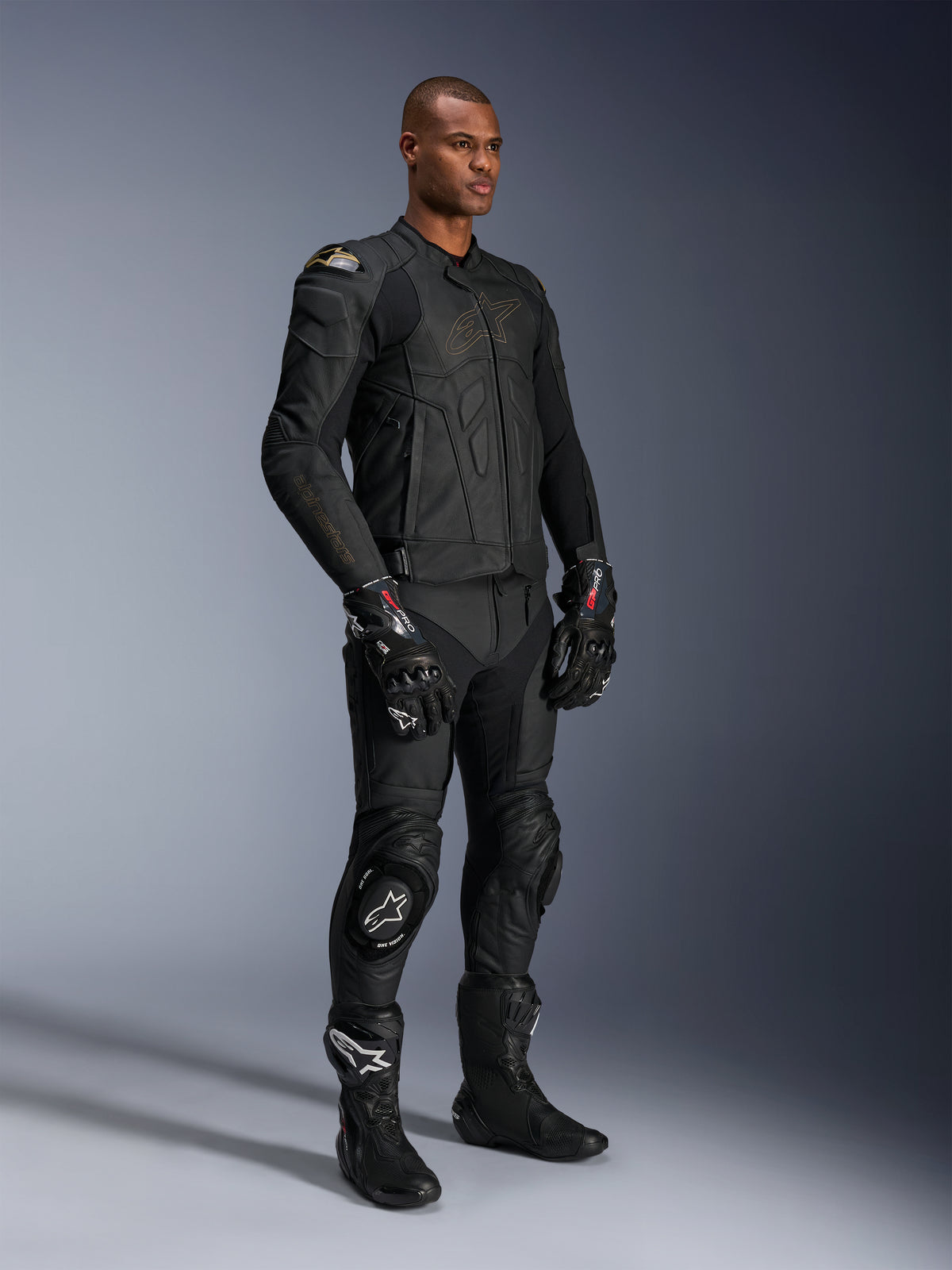 Modello che indossa pantaloni in pelle Alpinestars Missile V3 e giacca in pelle nera coordinata con dettagli oro, guanti racing GP Pro neri e stivali racing neri, equipaggiamento completo da moto per uso stradale, scatto in studio.