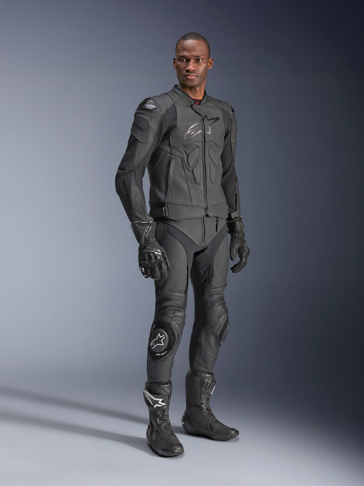 Giacca in pelle Alpinestars Dusk Airflow, Nero Nero, indossata da un pilota con equipaggiamento tecnico completo comprensivo di pantaloni in pelle, guanti e stivali coordinati, con pannelli in pelle traforata per la ventilazione e protezioni integrate per la guida sportiva