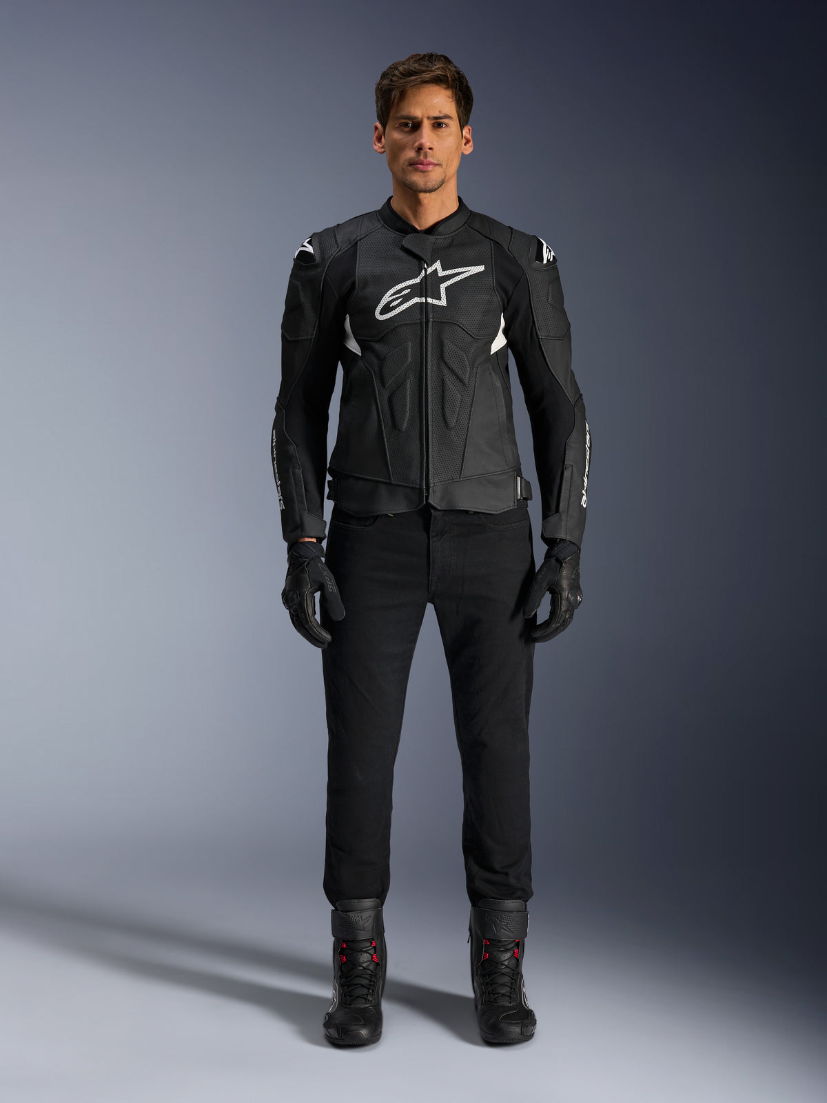 Alpinestars Dusk Airflow Leather Jacket, nero e bianco, giacca da moto in pelle con pannelli traforati per il flusso d'aria, logo Astars bianco sul petto, indossata da un pilota uomo con pantaloni neri, guanti e stivali da moto, scatto professionale in studio per equipaggiamento moto sportivo