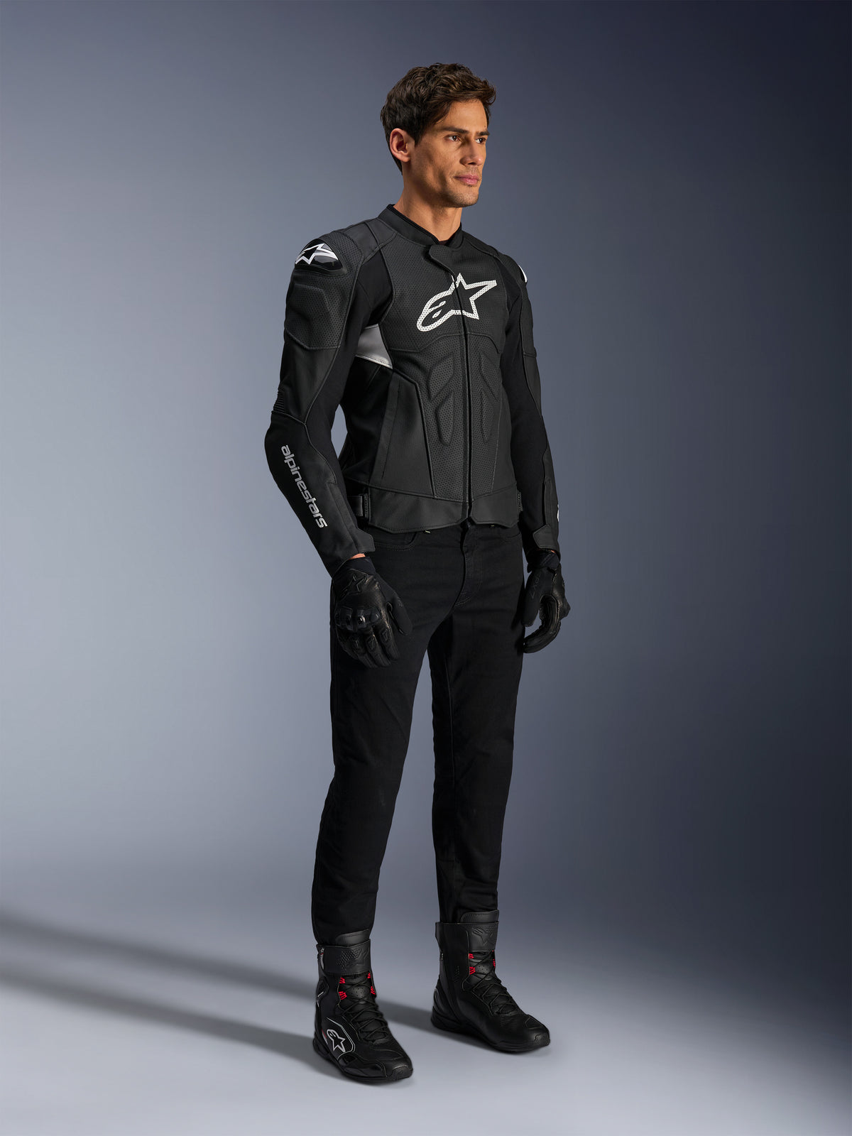 Alpinestars Dusk Airflow Leather Jacket, nero e bianco, indossata con pantaloni da moto, guanti e stivali neri, con pannelli in pelle traforata e loghi bianchi, stile racing sportivo.