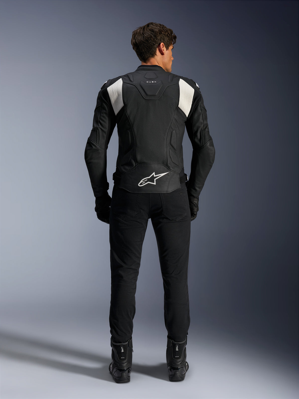 Giacca in pelle Alpinestars Dusk Airflow, nero e bianco, vista posteriore di un pilota con pantaloni e stivali neri, caratterizzata da pelle traforata per la ventilazione, pannelli a fisarmonica bianchi sulle spalle e logo bianco sulla parte bassa della schiena