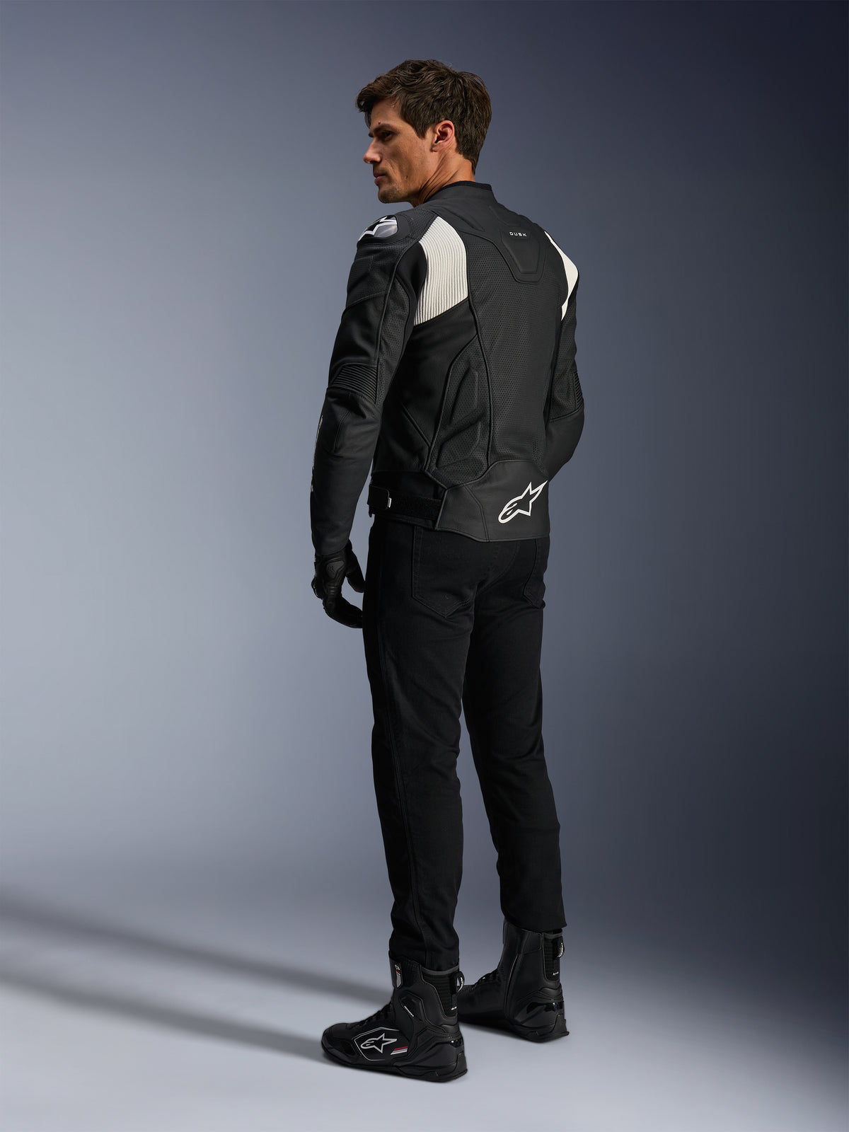 Giacca in pelle Alpinestars Dusk Airflow, nero e bianco, vista posteriore del modello che indossa la giacca in pelle traforata con pantaloni e stivali neri coordinati, caratterizzata da pannelli elastici a fisarmonica bianchi e logo sulla parte bassa della schiena, abbigliamento tecnico da moto