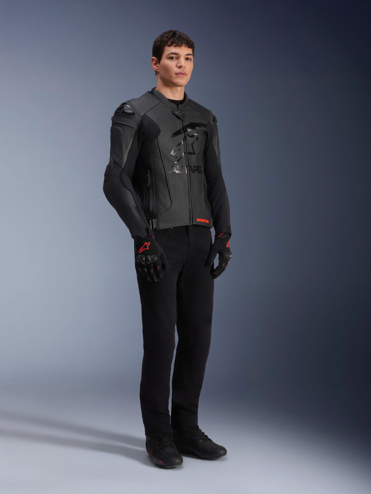 Giacca in pelle Alpinestars GP Plus R V4 Airflow, nero, indossata da un modello uomo con pantaloni da moto neri e guanti con dettagli rossi, caratterizzata da pannelli in pelle traforata e slider sulle spalle integrati, scatto in studio a figura intera.