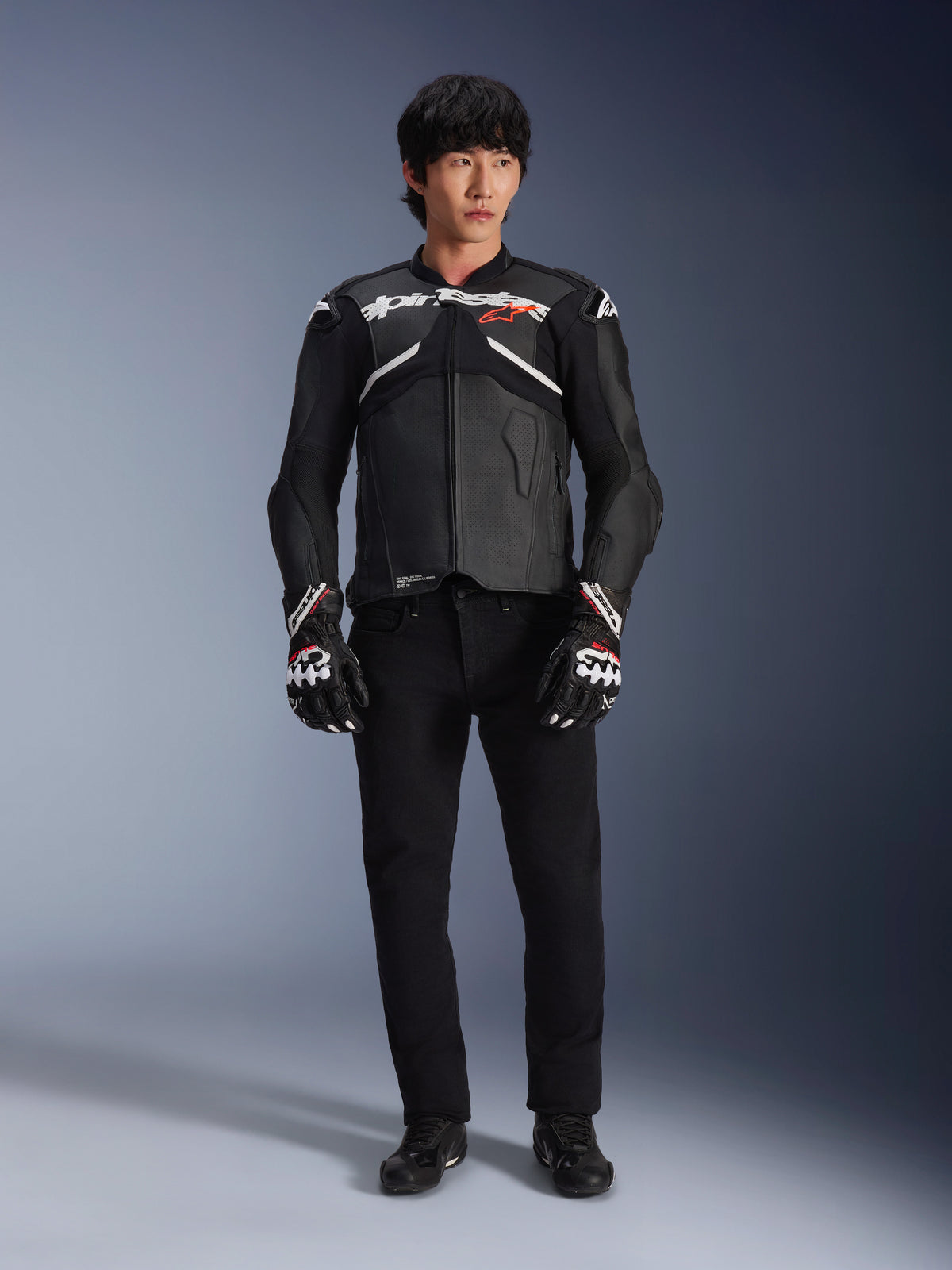 Modello che indossa la giacca in pelle Alpinestars Atem V5 in nero e bianco, con marchio Alpinestars bianco e logo con stella rossa sul petto, abbinata a pantaloni neri e guanti da corsa bianchi e neri, abbigliamento da moto performante ed elegante