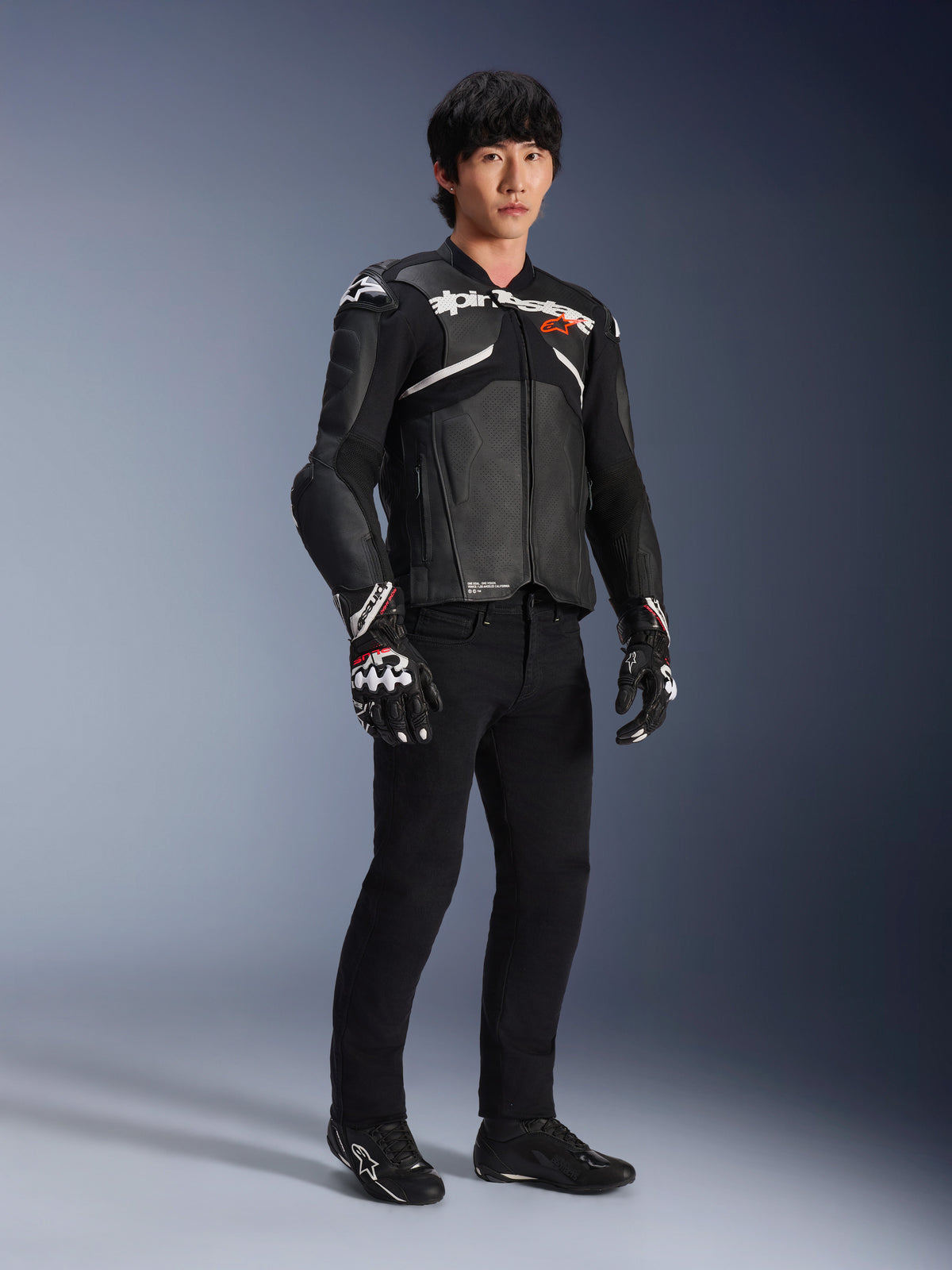 Giacca in pelle Alpinestars Atem V5, giacca da moto racing, nero e bianco, indossata con pantaloni neri e guanti coordinati, con logo Alpinestars bianco sul petto e protezioni sulle spalle
