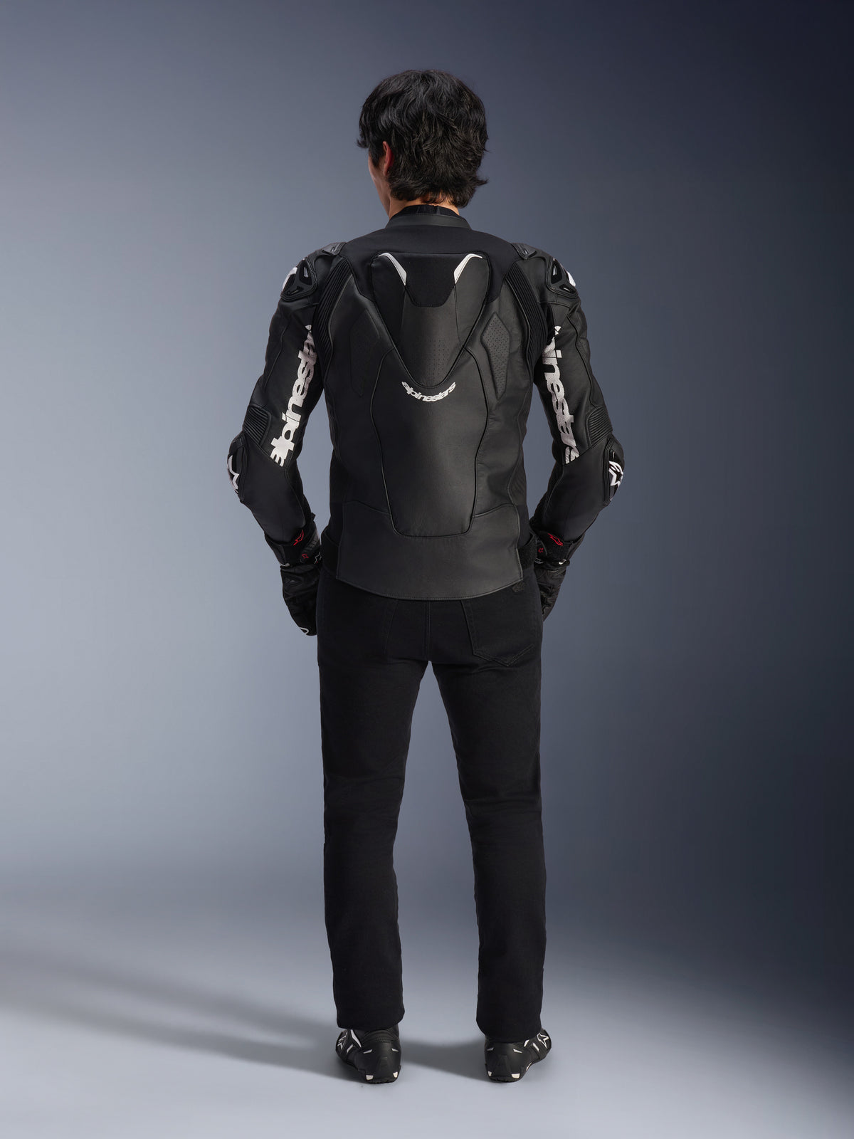 Giacca in pelle Alpinestars Atem V5, giacca da moto racing, nero e bianco, vista posteriore sul pilota, dotata di gobba aerodinamica e branding Alpinestars bianco sulle maniche e sulla schiena, struttura in pelle tecnica, indossata con pantaloni neri e stivali da moto