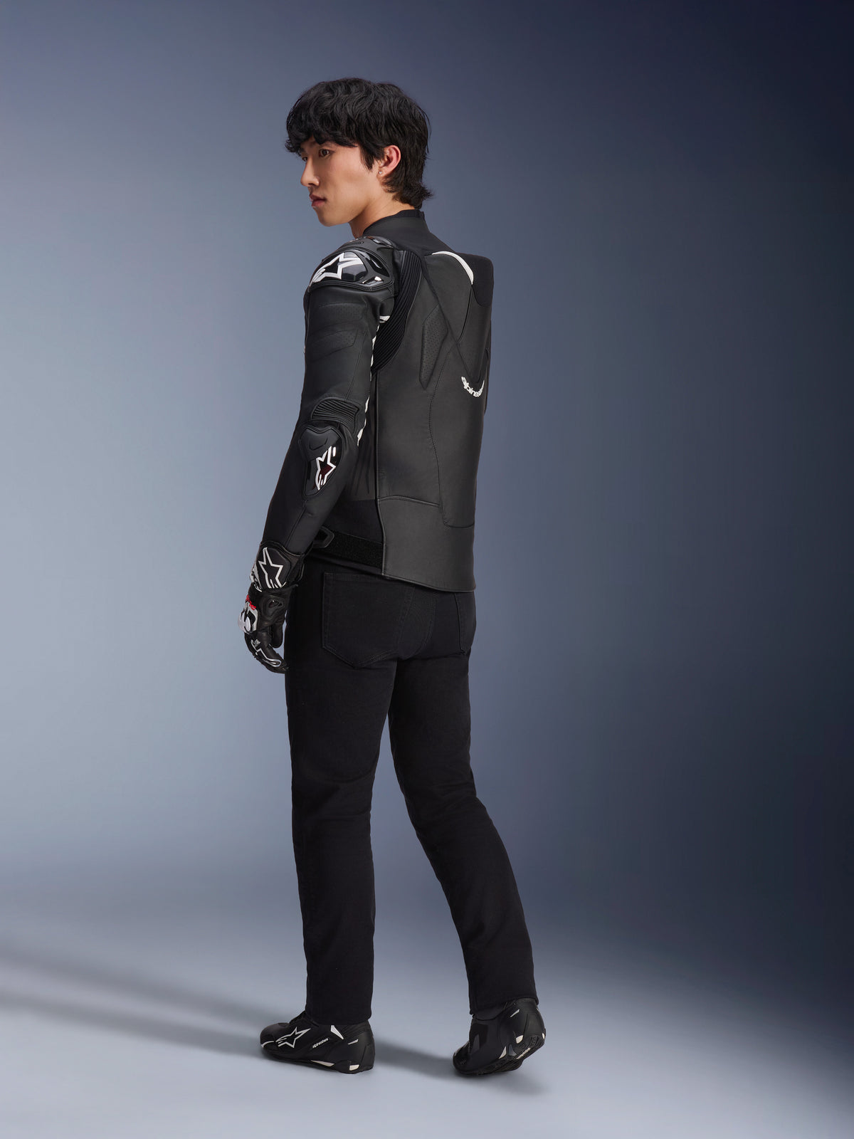 Modello che indossa la giacca in pelle Alpinestars Atem V5 in nero e bianco, vista posteriore a tre quarti che mostra la gobba aerodinamica e le protezioni DFS RR, abbinata a pantaloni neri, guanti racing e stivali, abbigliamento professionale da moto