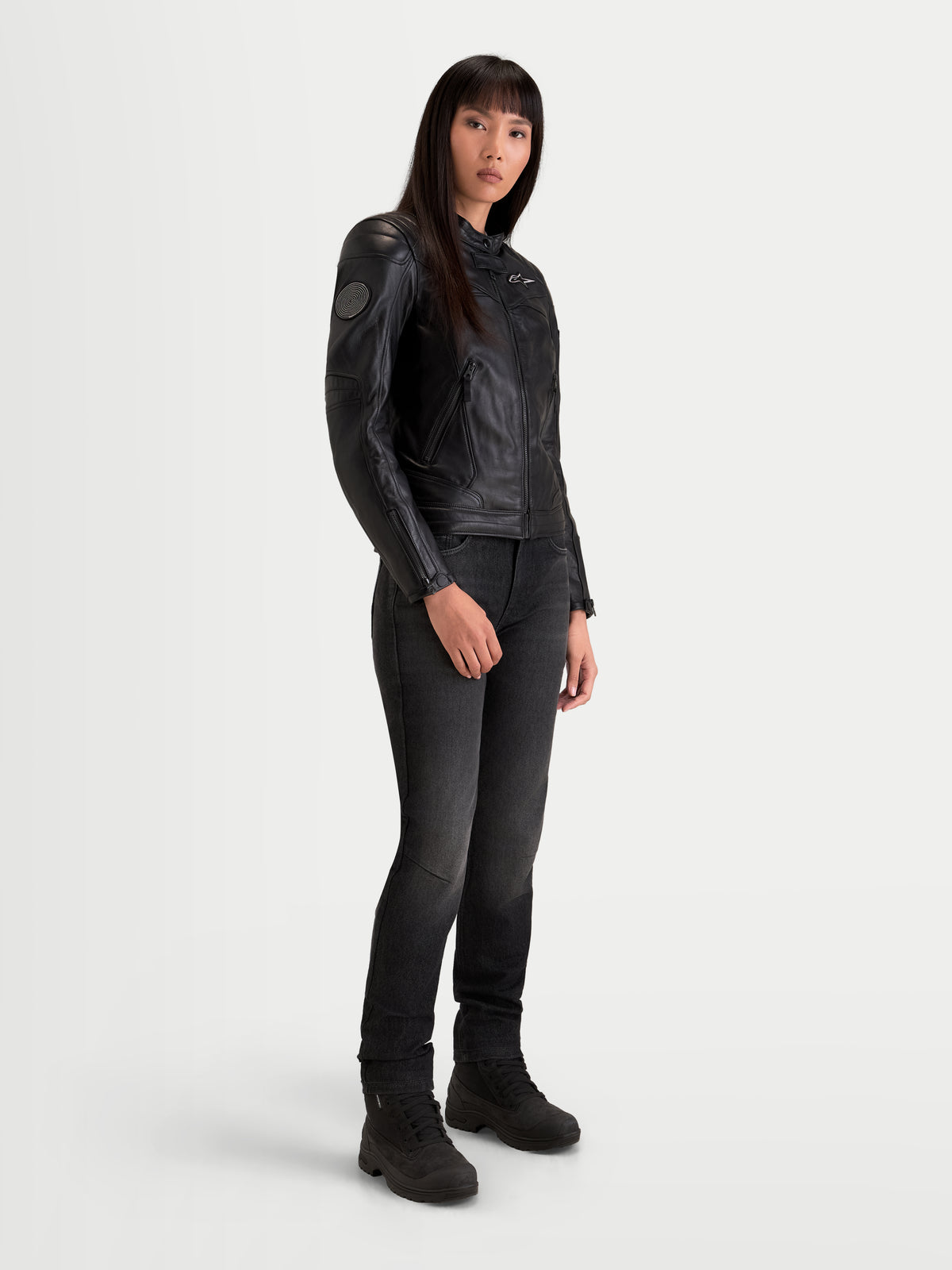 Alpinestars Revea Donna Giacca di pelle, giacca da moto urbana contemporanea da donna in nero, vista tre quarti su una modella che indossa jeans scuri e stivali da moto, struttura in pelle bovina pieno fiore con certificazione di sicurezza AA